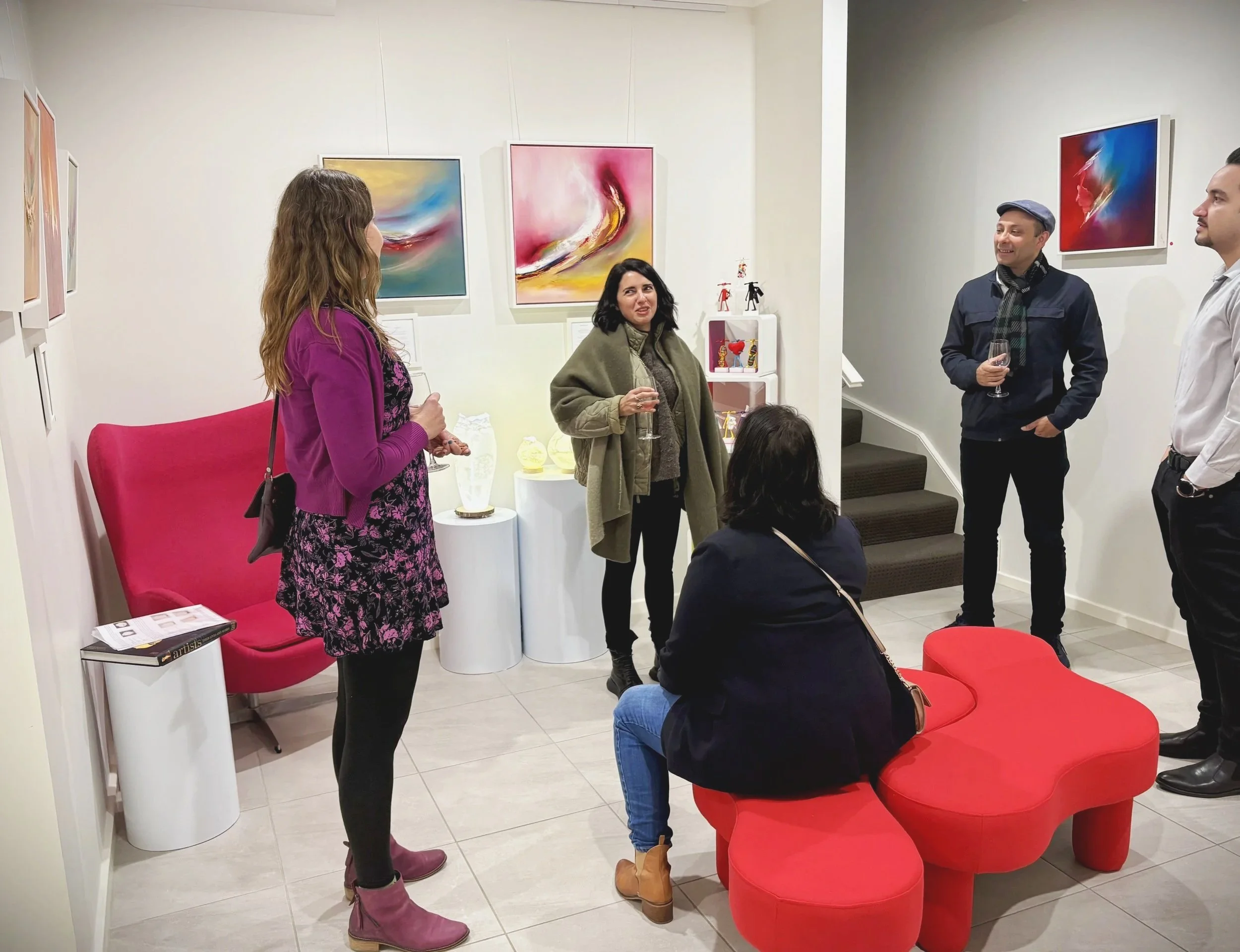 Group of five people socializing in an art gallery with colorful abstract paintings on the walls. Two women are seated on a red, kidney-shaped bench, while three standing are holding wine glasses and engaging in conversation. Mira Corbova Art Gallery
