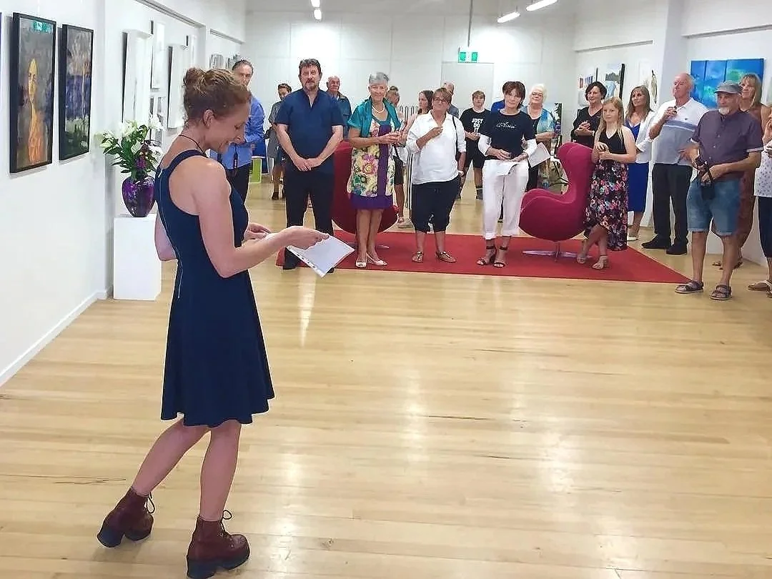 A woman in a blue dress, an artist speach at the art exhibition at the gallery with a big crowd. 