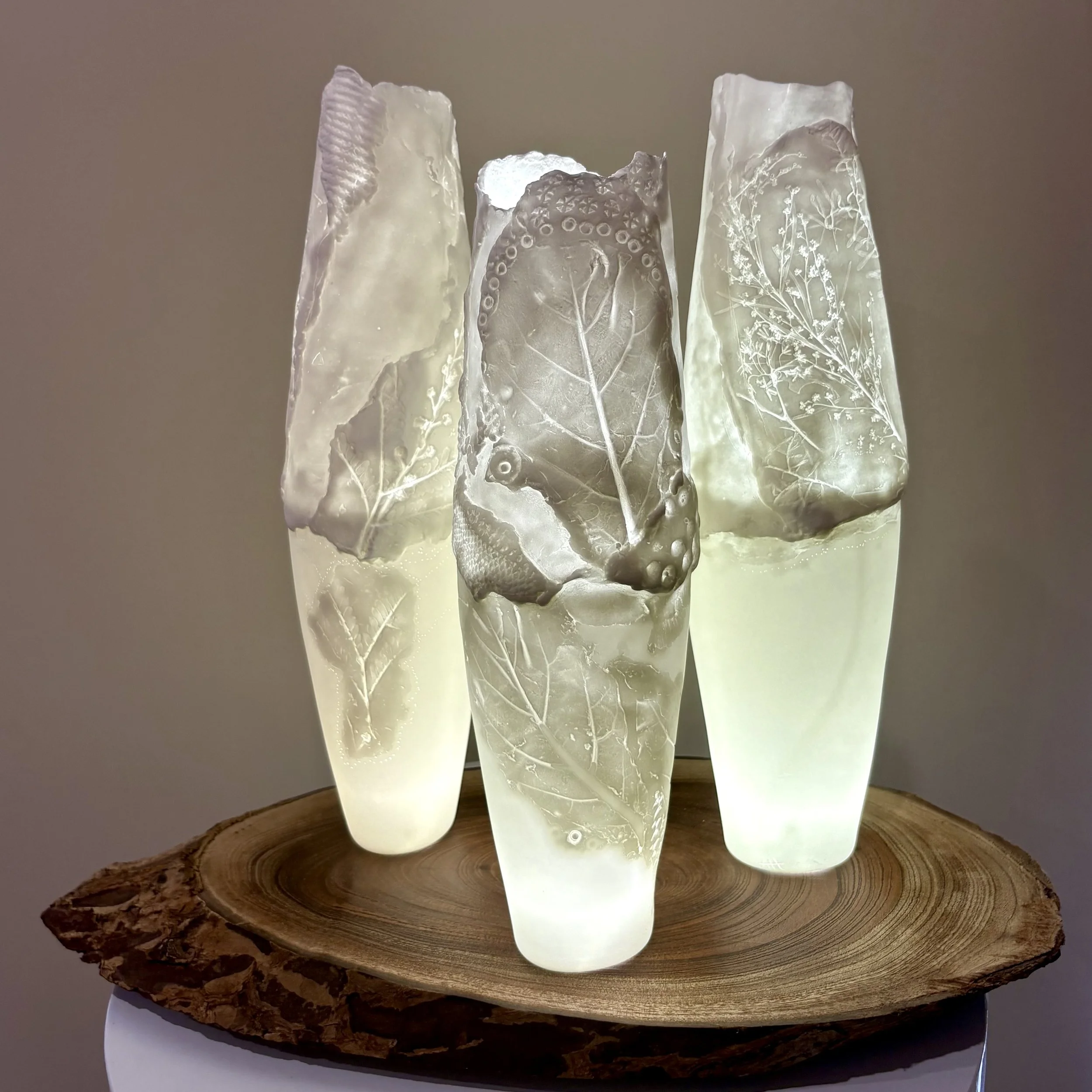 Three tall glass vases with etched leaf and floral designs, placed on a wooden slab, illuminated with soft light.
