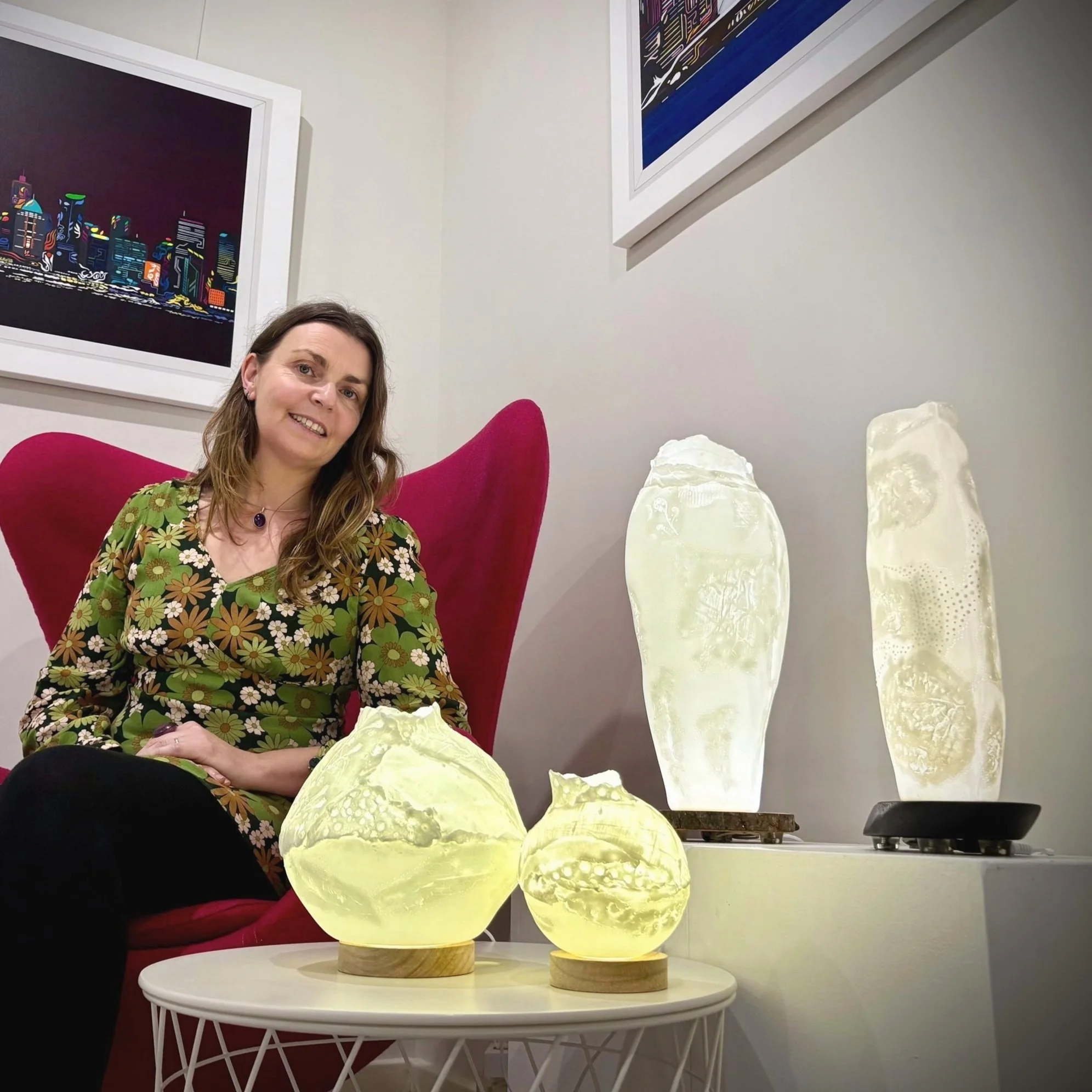 A woman with long brown hair sitting in a pink chair, wearing a colorful floral dress, smiling at the camera. In front of her on a white table, there are four glowing, abstract sculptures resembling rough spheres and elongated shapes, illuminated with a soft yellowish light. Behind her are two tall illuminated sculptures on a white surface and two framed artworks on the wall.
