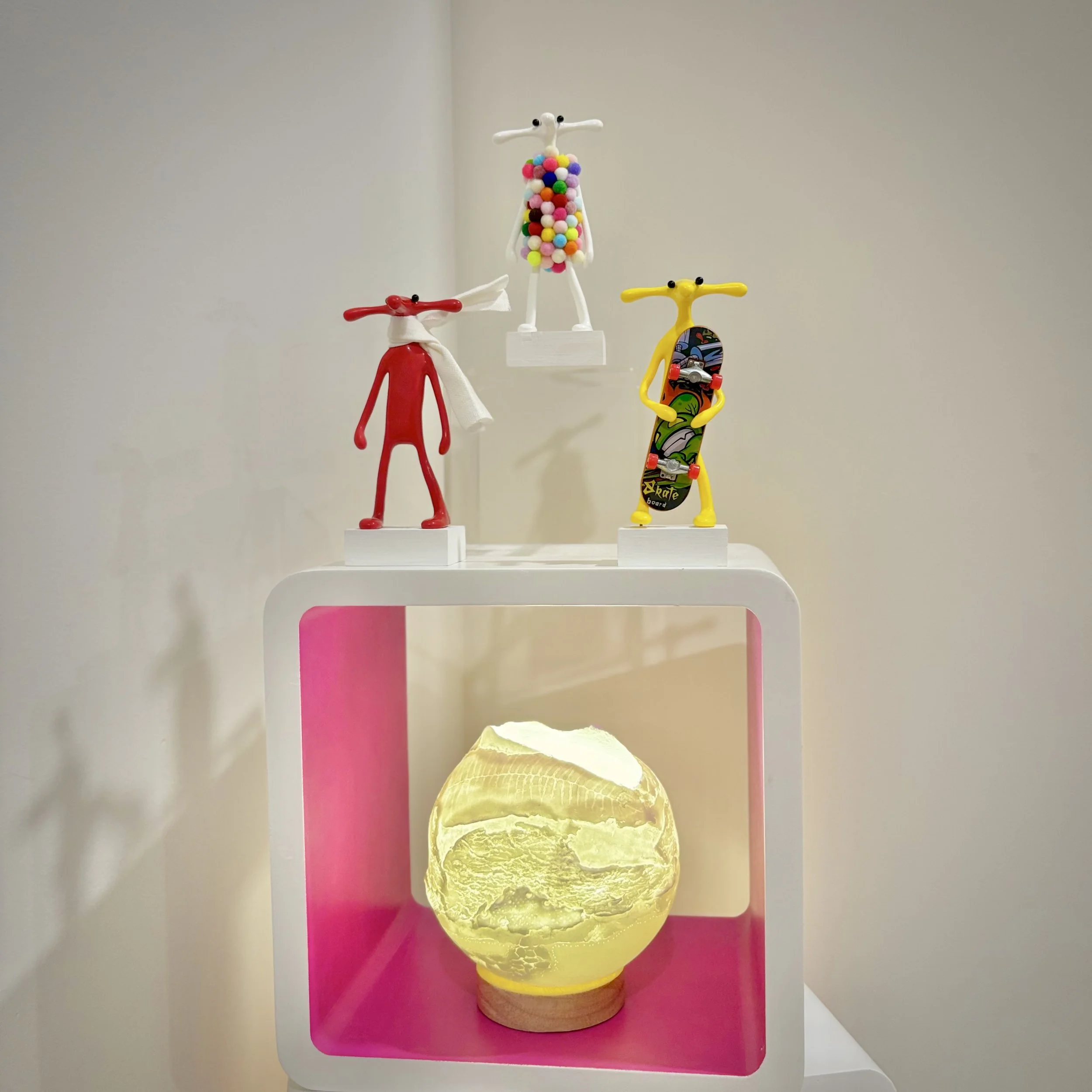 Three colorful figurines resembling anthropomorphic dogs on white display stands, with a yellow-lit decorative orb on a pink shelf below.
