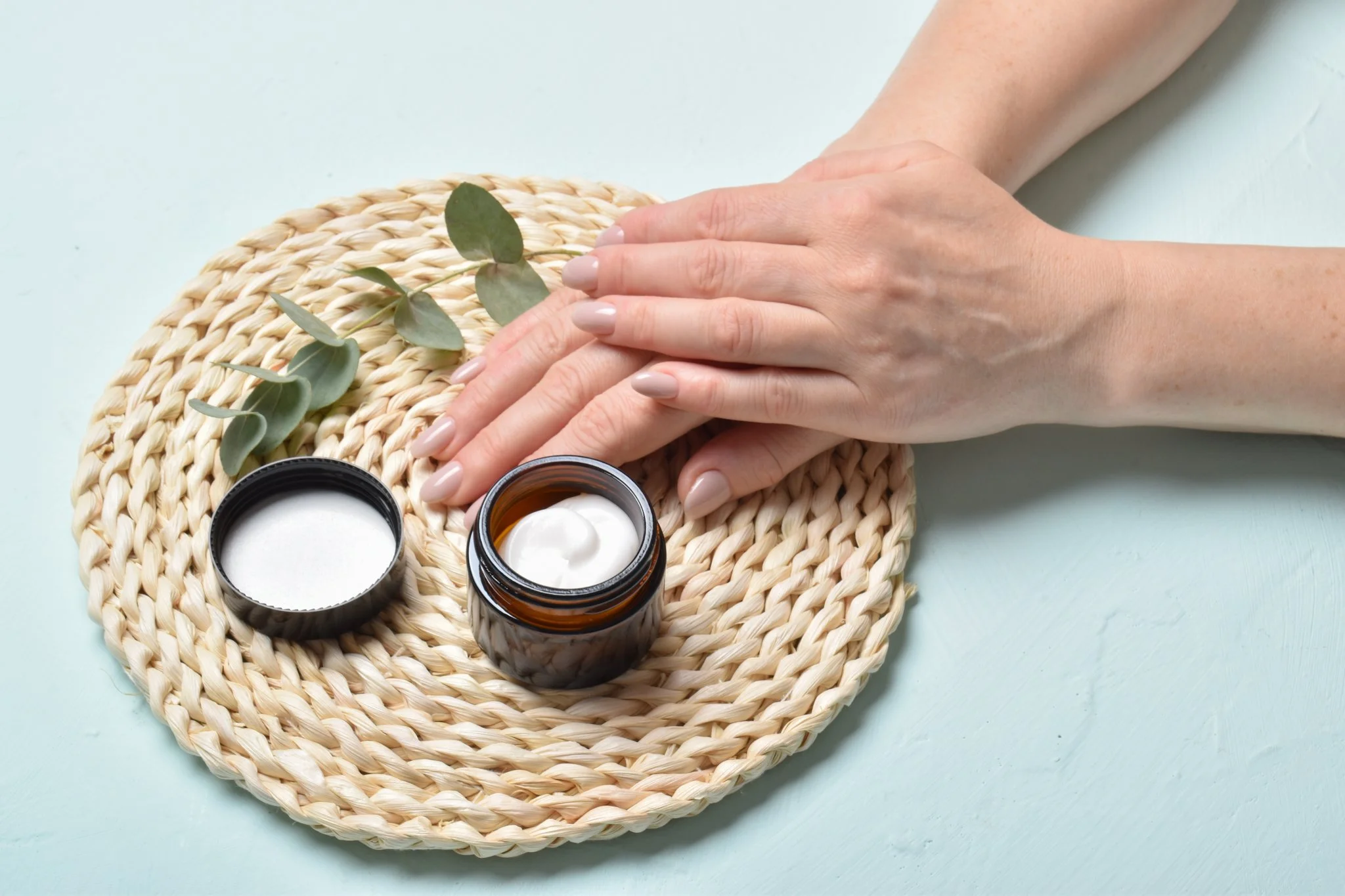Hands with manicured nails resting over a woven placemat, with an open jar of cream, a container with white powder, and eucalyptus leaves on a light-colored surface.
