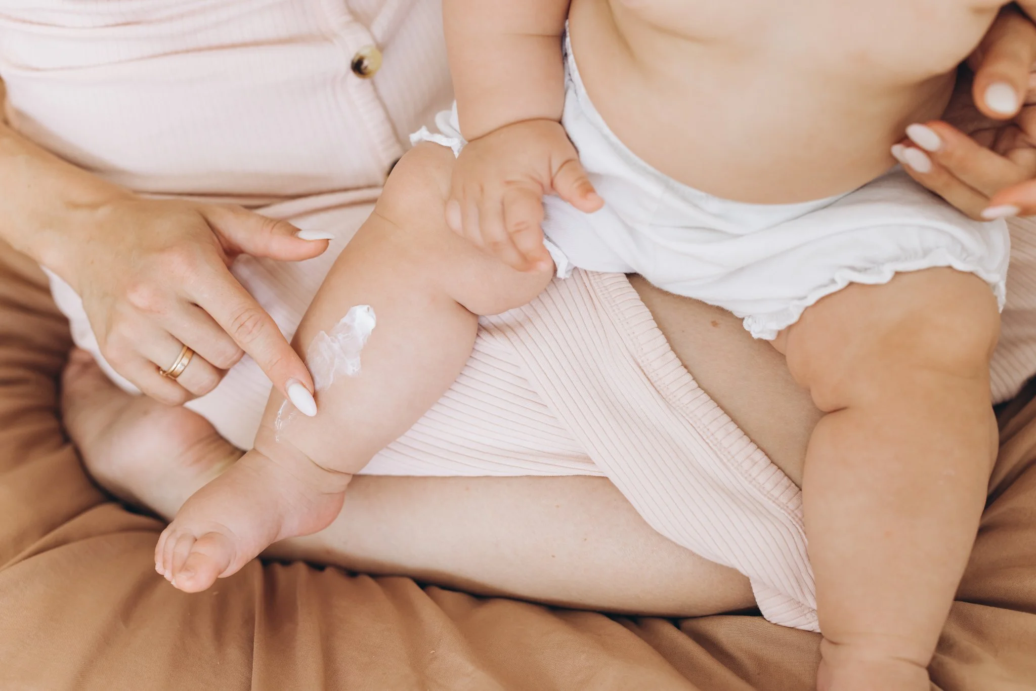 A person applies lotion to a baby's leg while holding the baby's other leg. The baby is sitting on the person's lap, wearing a diaper, and the person is wearing a pink top.