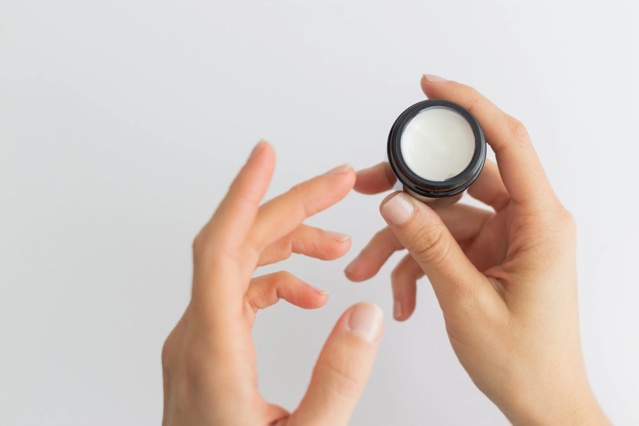 A person holding a small black jar of white cream or lotion against a plain white background.