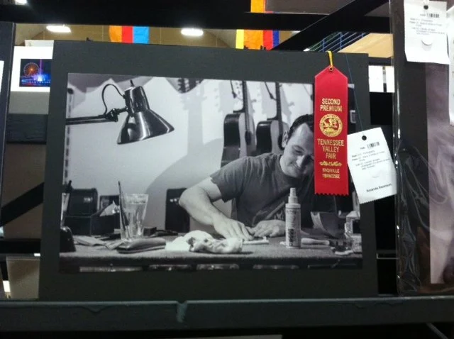 Black and white photo of a man sitting at a desk, writing, with a lamp, a glass, and various items on the desk.