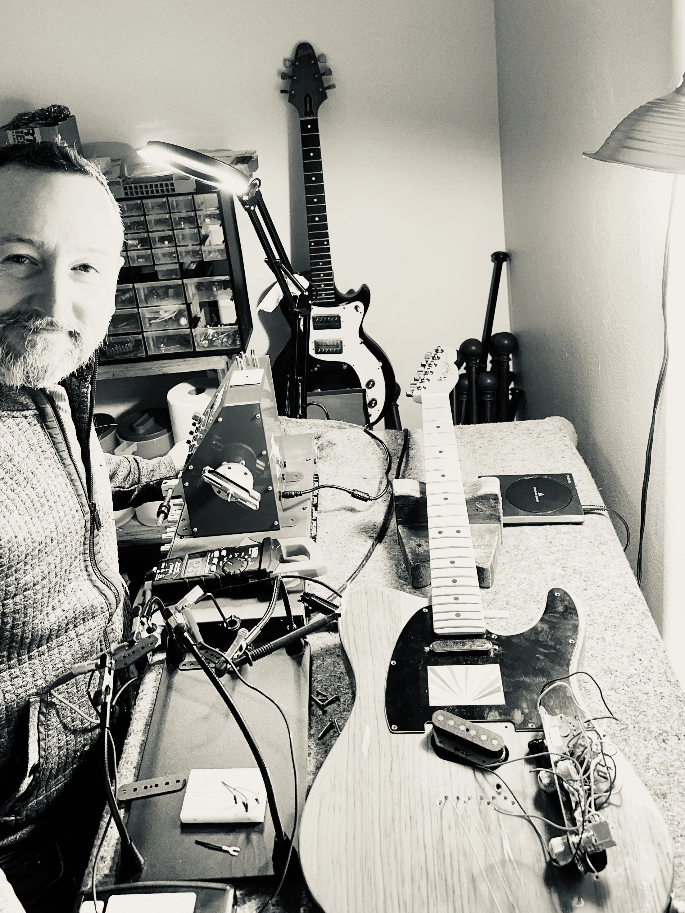 A man working on assembling an electric guitar in a home workshop, with various guitar parts, tools, and electronic equipment on the workbench, and a finished guitar hanging on the wall.