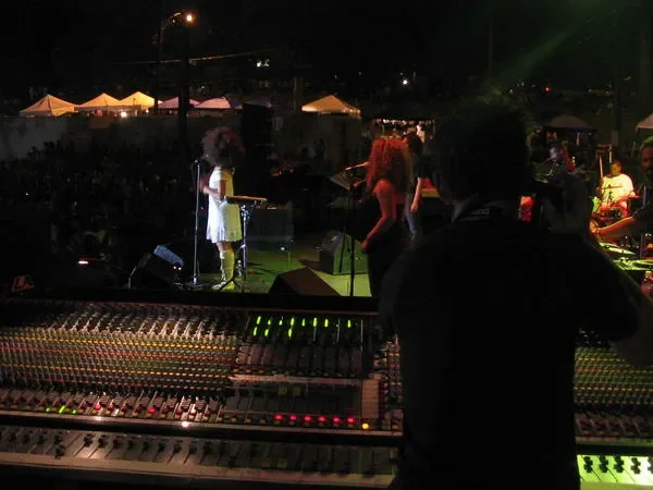 A woman in a white dress performing on a stage at night, with band members and sound engineers, colorful tents and lights in the background.