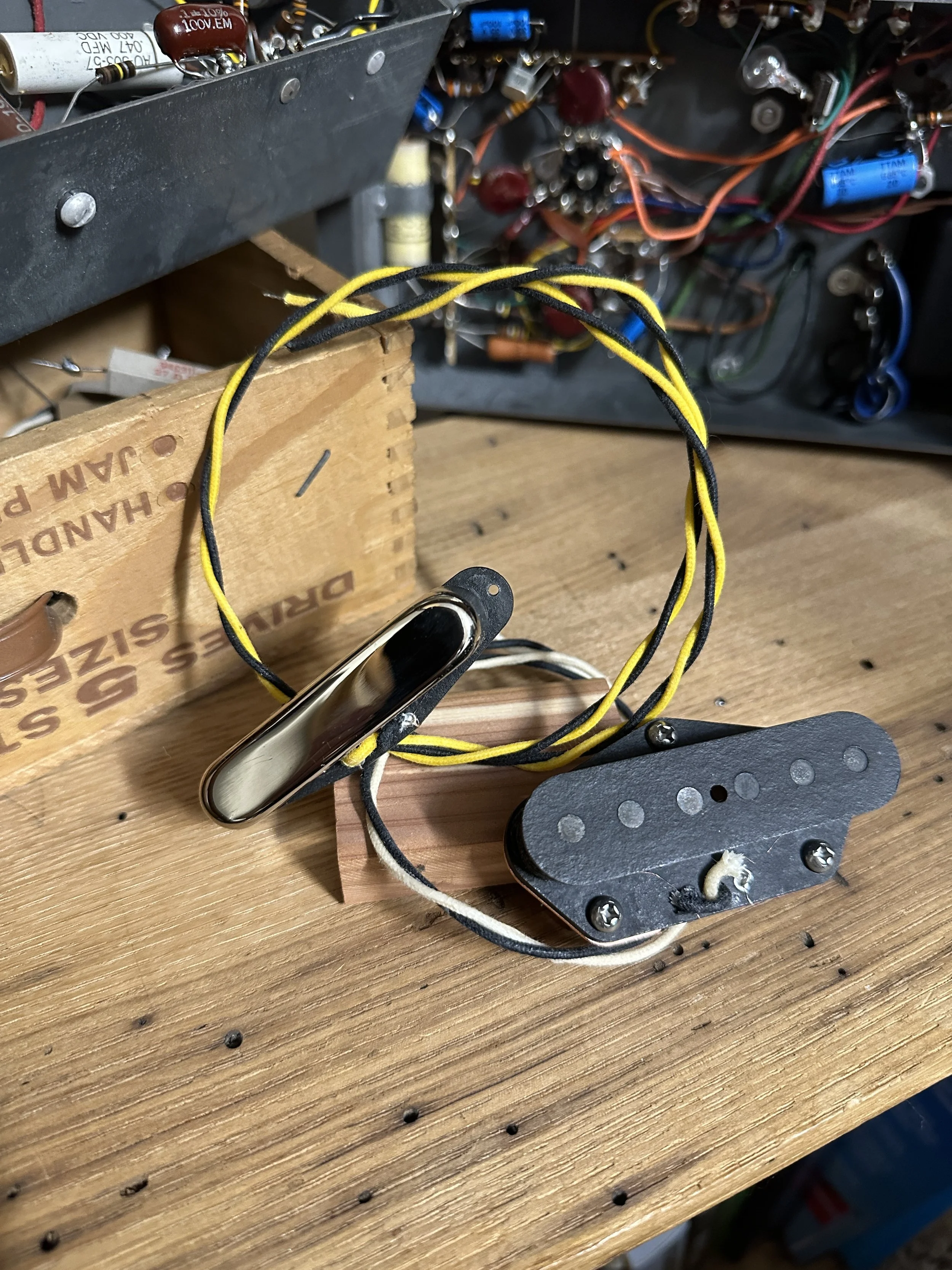 An electric guitar pickup with wires and a metal cover, placed on a wooden workbench with electronic components and wiring in the background.