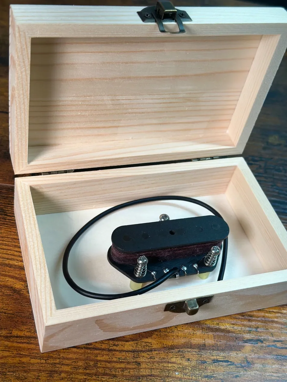 A wooden box with a latch, inside of which is a black electric guitar pickup with metal springs and wires.