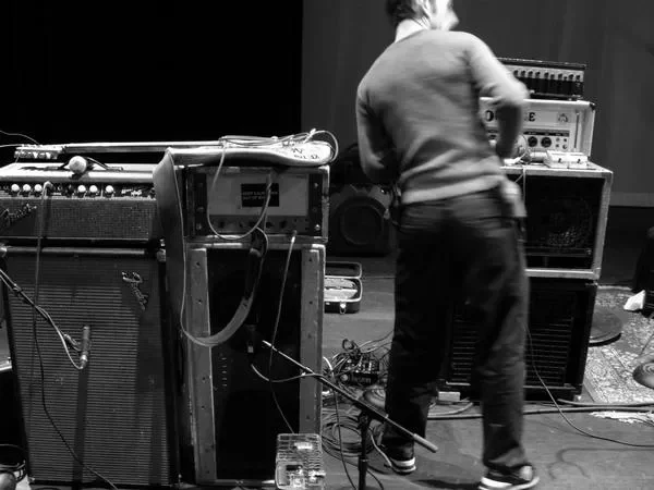 A person adjusting sound equipment in a music studio or rehearsal space with various amplifiers, speakers, and audio gear.