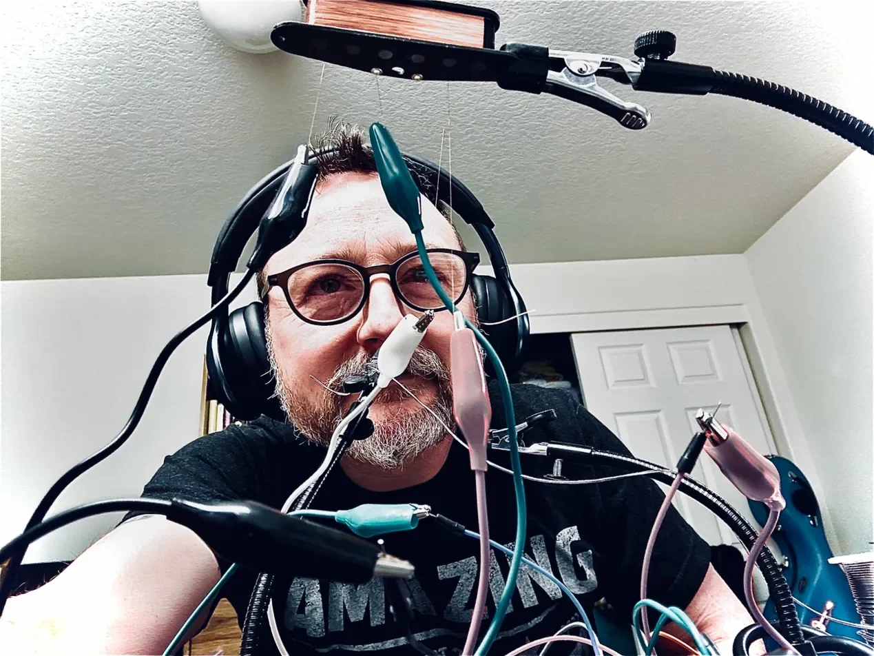 Man with glasses and headphones working on electronics with wires and tools in a room.