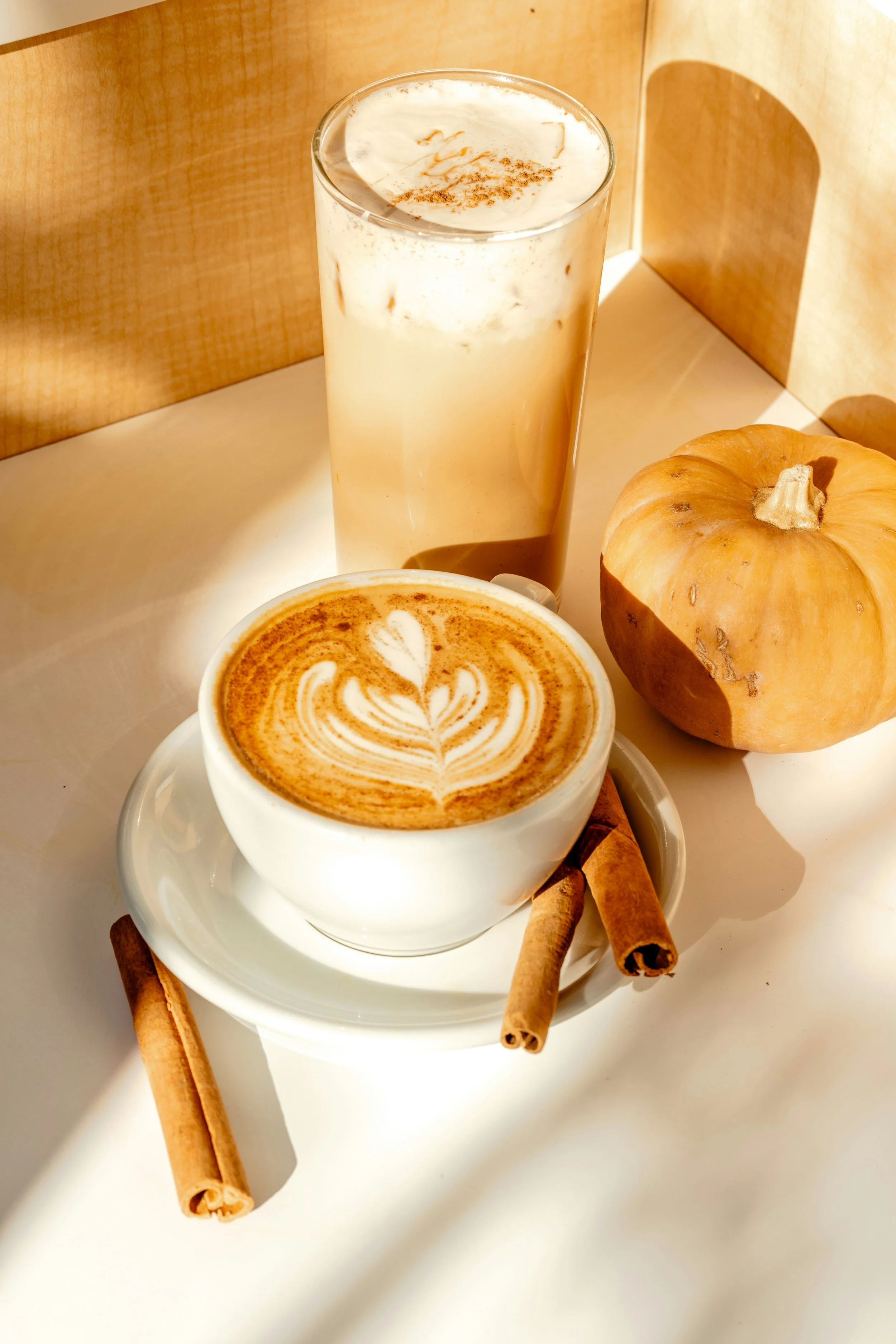 A latte with latte art, a cinnamon stick, a pumpkin, and an iced coffee on a white table.