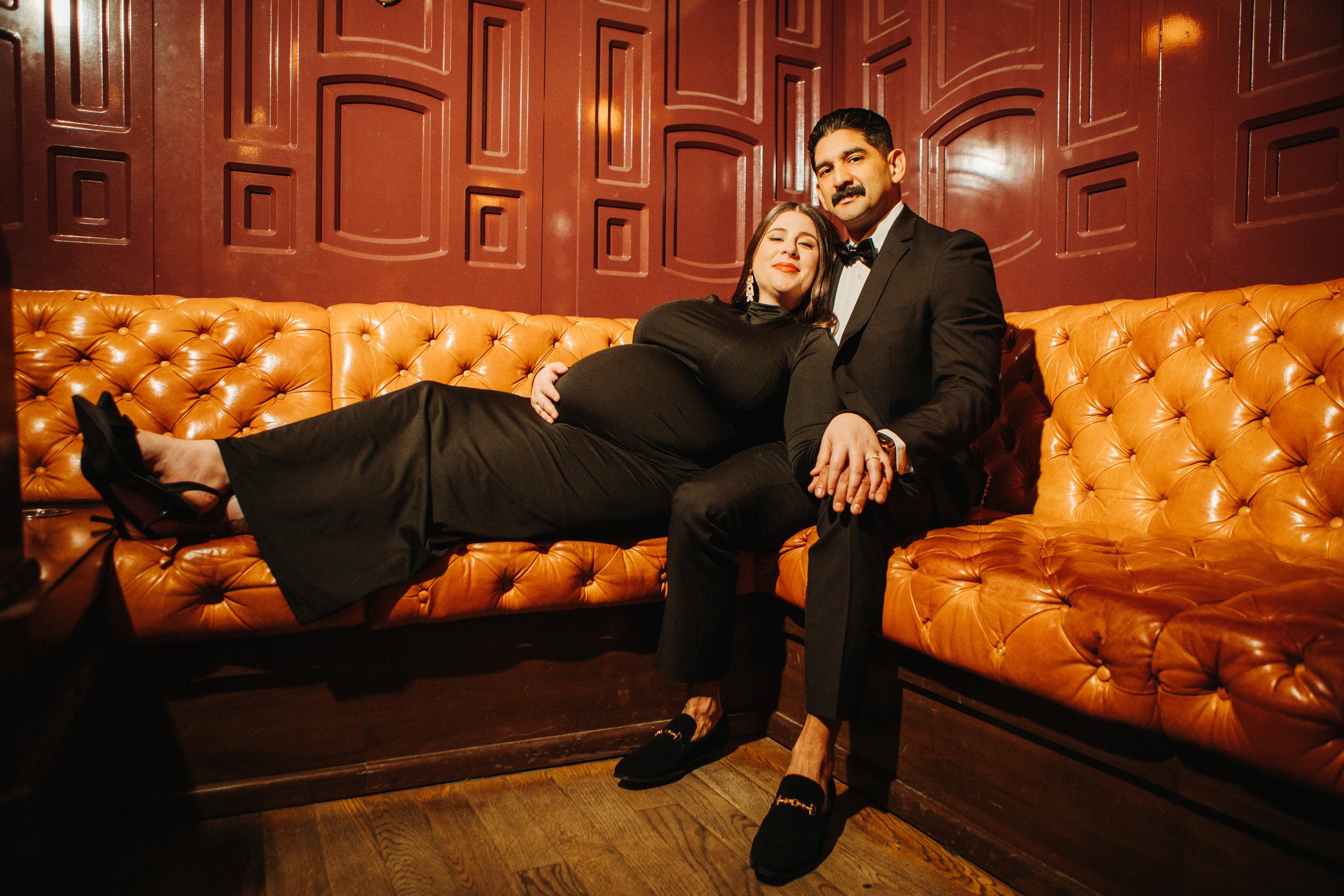 An elegant maternity portrait of a couple reclining on a tufted caramel leather banquette at the famous Lafayette Hotel in San Diego. The expectant mother wears a sleek black dress and rests comfortably against her partner, who is dressed in a black 