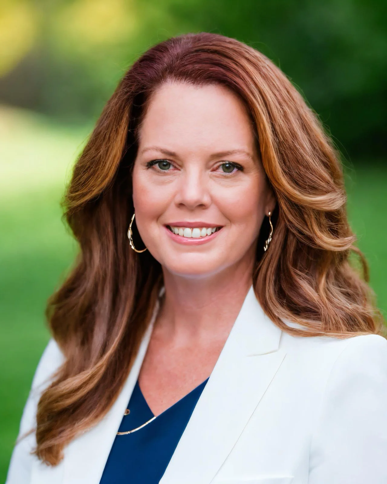A smiling woman with long auburn hair wears a white blazer over a navy top, posed outdoors against a softly blurred green background.