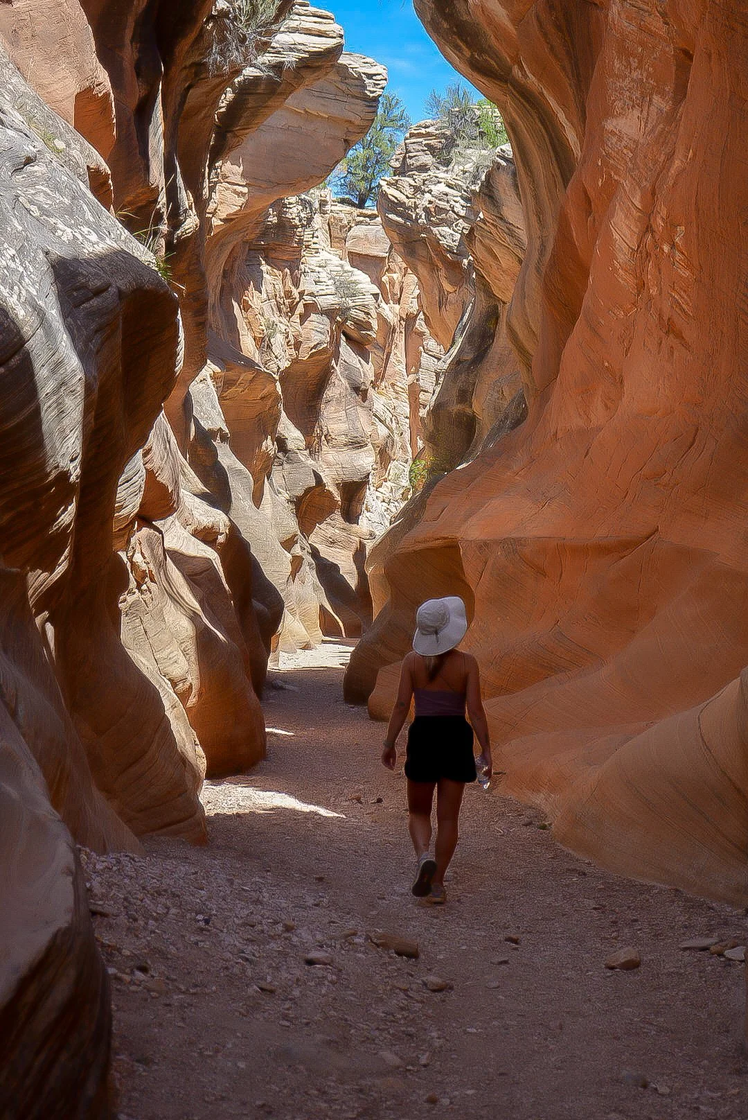 A person walking through a narrow, winding canyon with tall, colorful sandstone walls and a bright blue sky overhead.