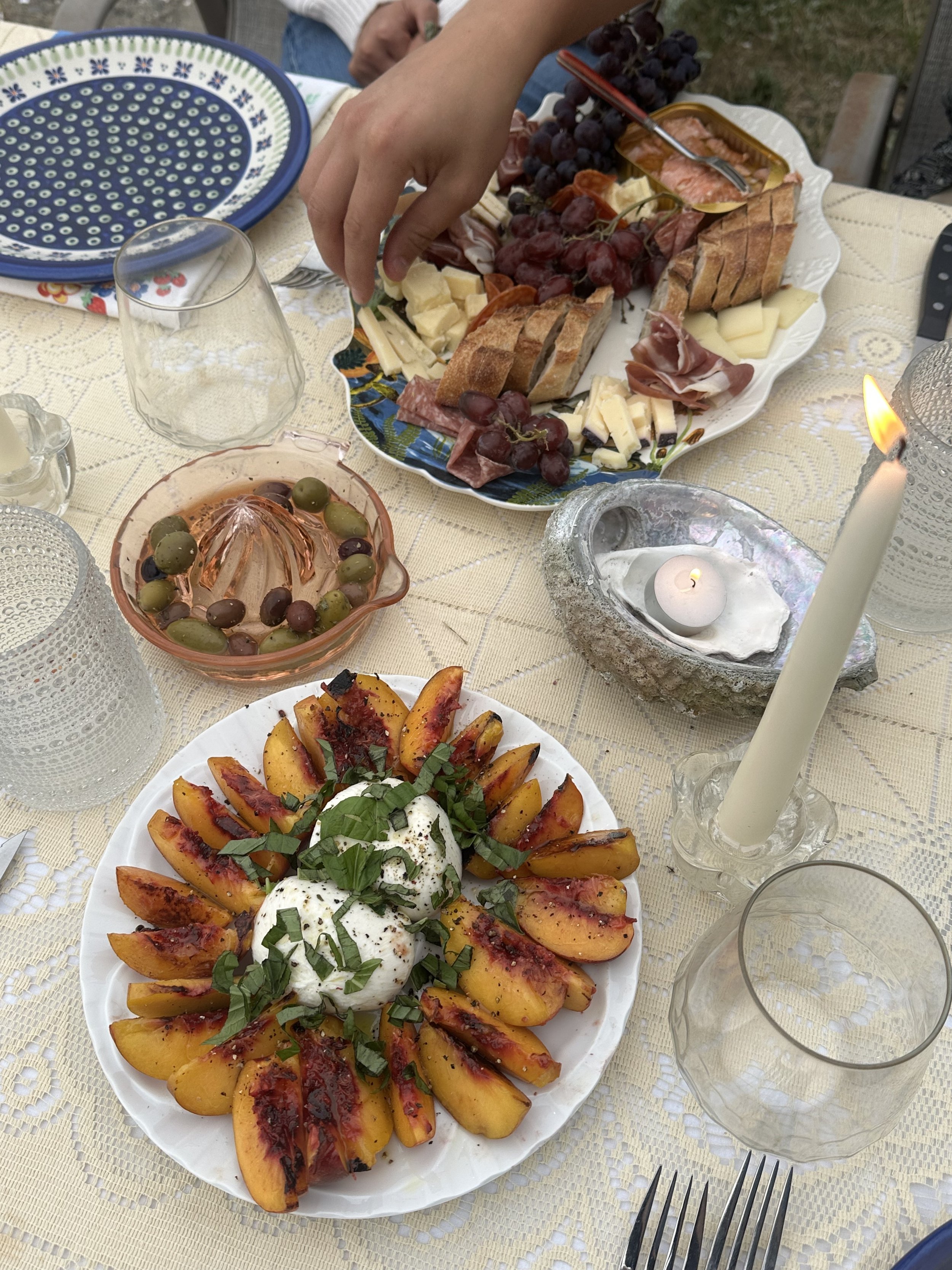 A table set with various dishes including a plate of sliced peaches topped with herbs and a white creamy topping, a bowl of mixed green and purple olives, and a large platter of assorted cheeses, meats, grapes, and bread. Candles and empty glasses are also on the table.