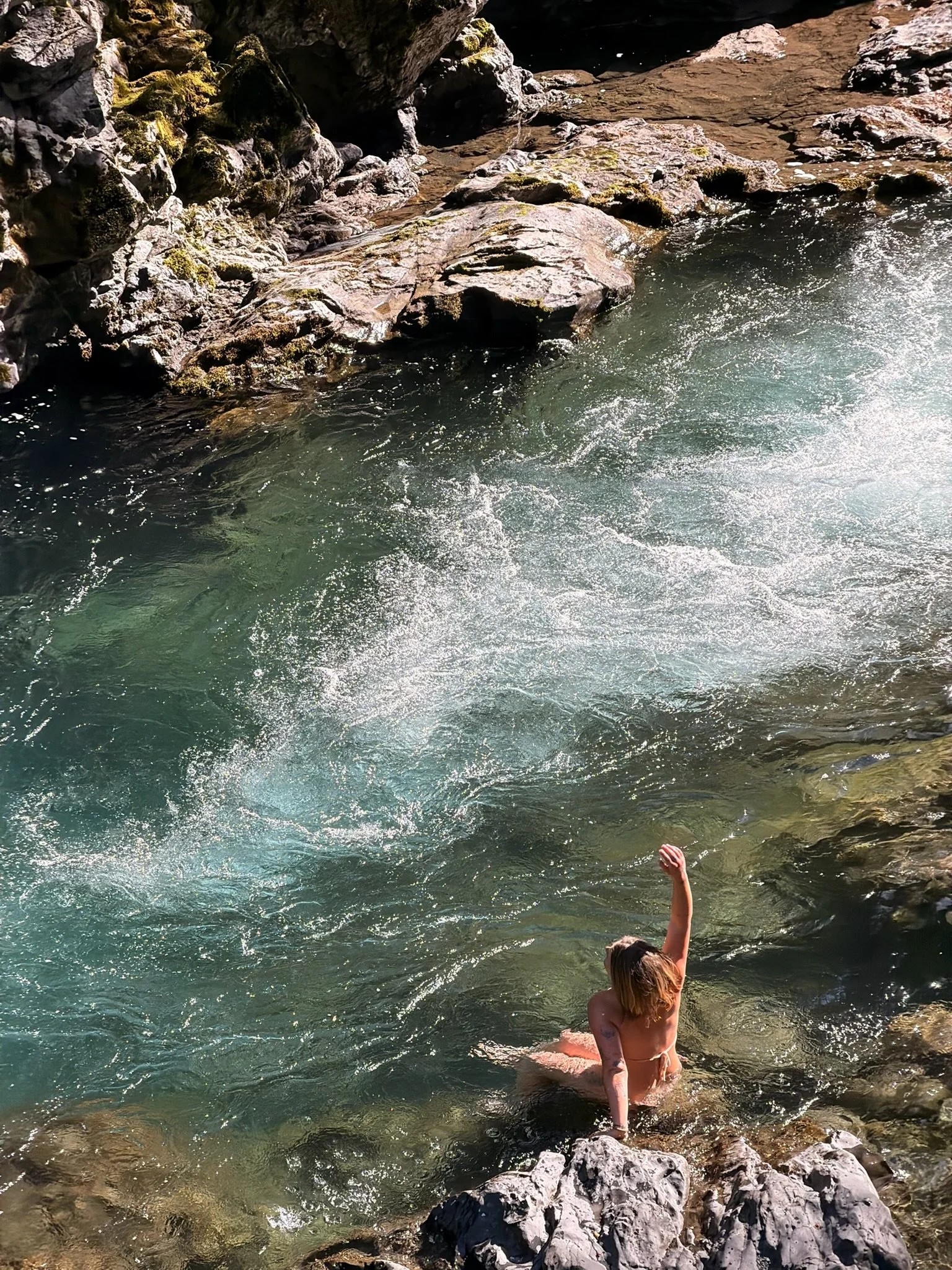 A person with long hair is sitting on rocks at the edge of a river or stream, with one arm raised and water splashing around. The scene includes rocks, water, and natural surroundings.