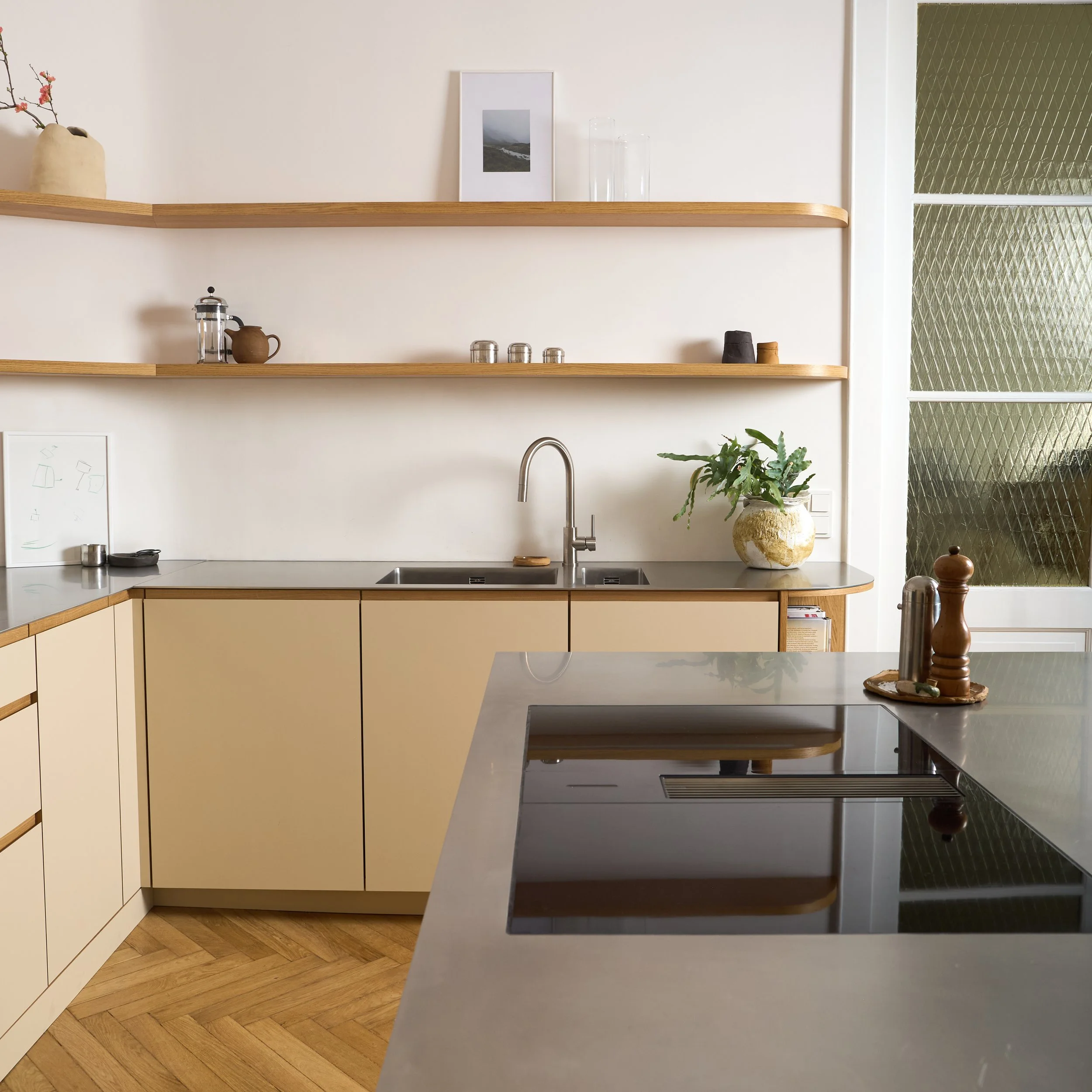 Moderne, minimalistische Küche mit beige Schränken, Holzregalen, Edelstahl-Spüle, Topf und Pflanzen auf dem Fensterbrett, Eichenparkett, Glastür mit Muster, Kochfeld auf Steinplatte, dekorativen Gewürzstreuern und einem Bild auf der Wand.