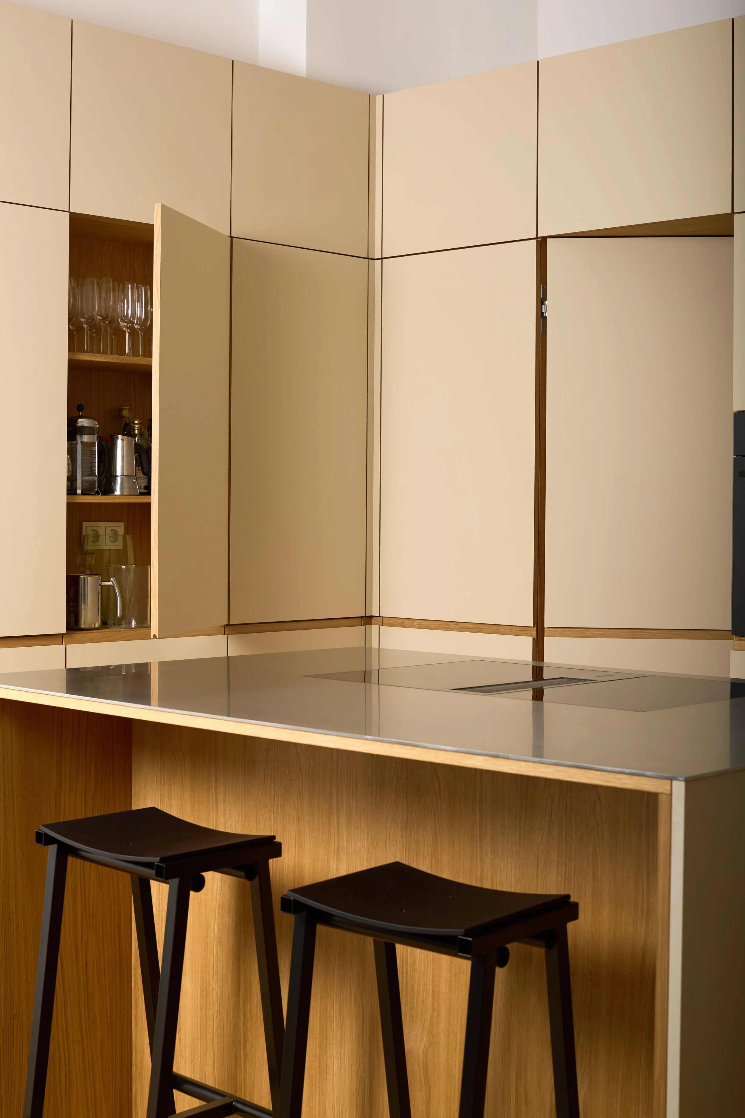 Moderner Küchenbereich mit dunkelbraunen Barhockern, einer schwarzen Herdplatte und beige Schränken mit Holzdetails.