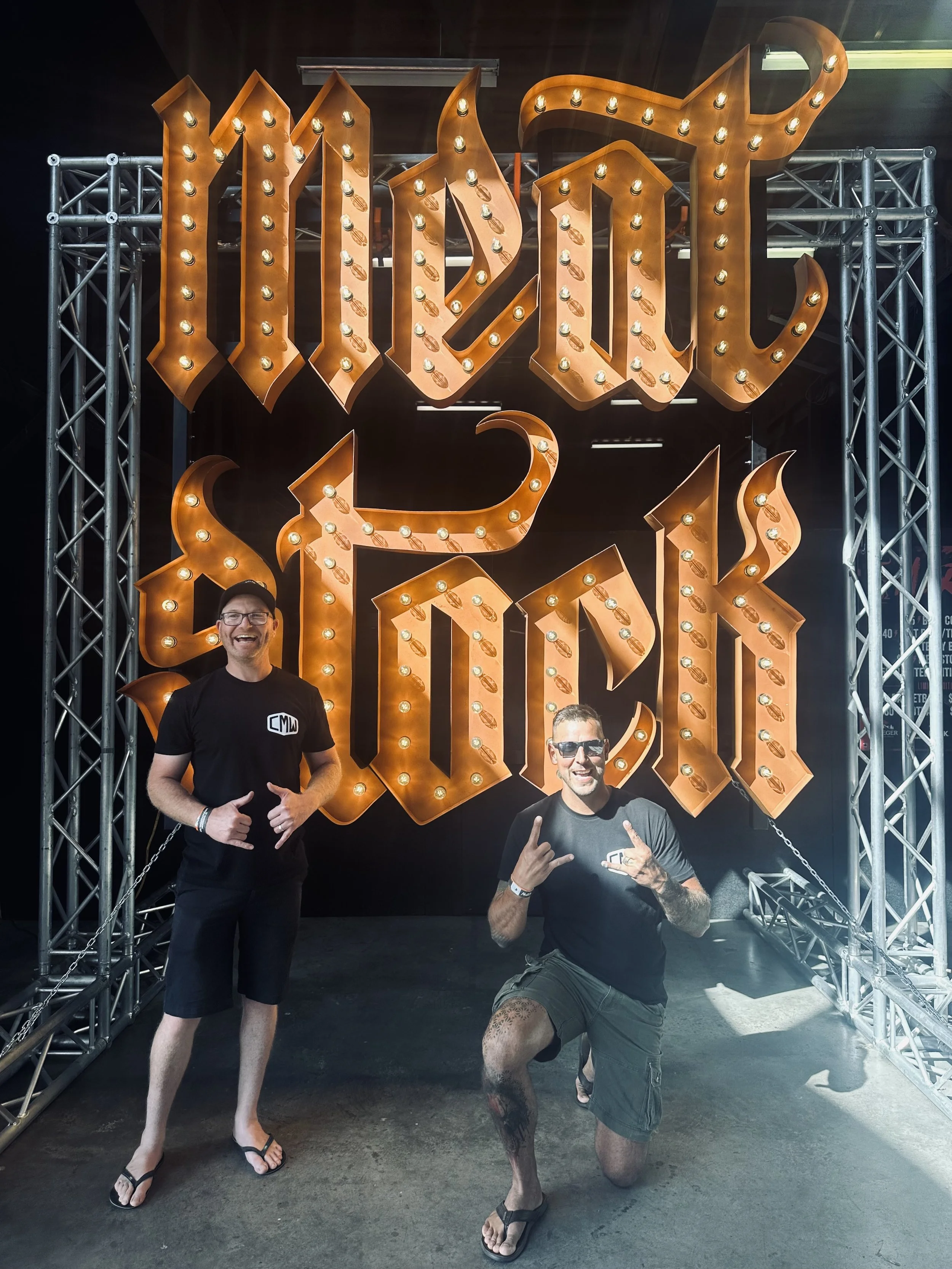 Two people posing in front of a large, illuminated "Meat Stock" sign, wearing casual clothing and flip-flops.