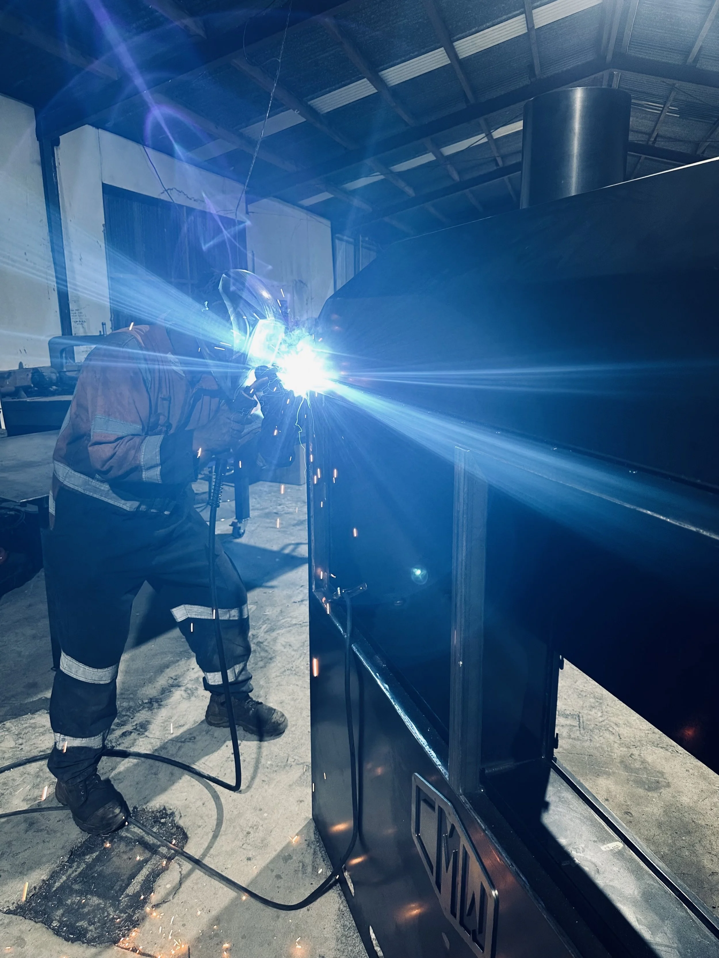 A person welding metal in an industrial workshop, wearing protective gear with bright sparks and light emanating from the welding process.