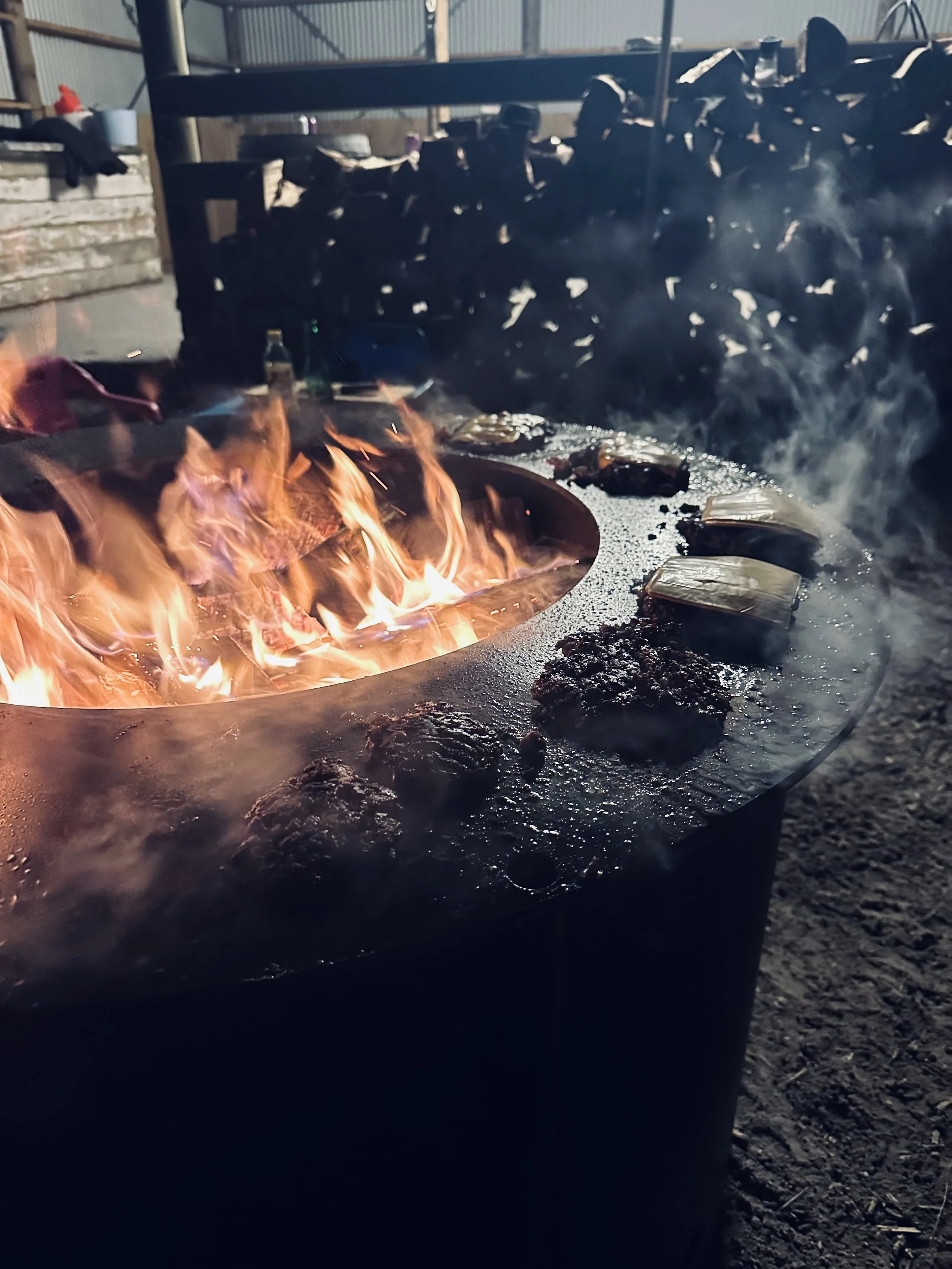 Outdoor fire pit with burgers and cheese on grill, surrounded by smoke, under a shelter with woodpile in background.