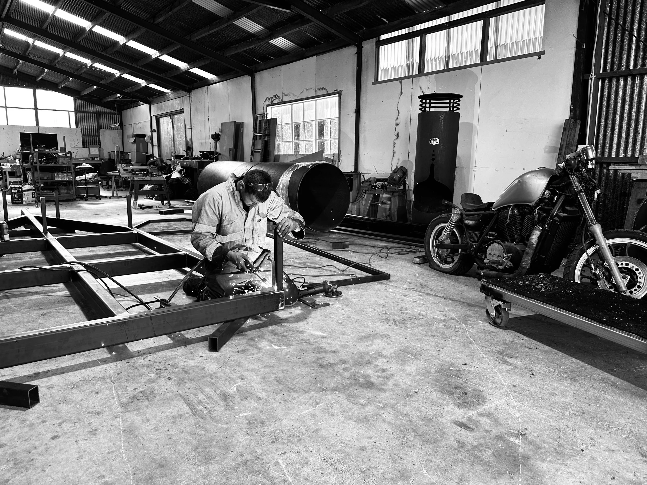 Black and white image of a welder in a workshop working on a metal frame, with a motorcycle and industrial equipment in the background.