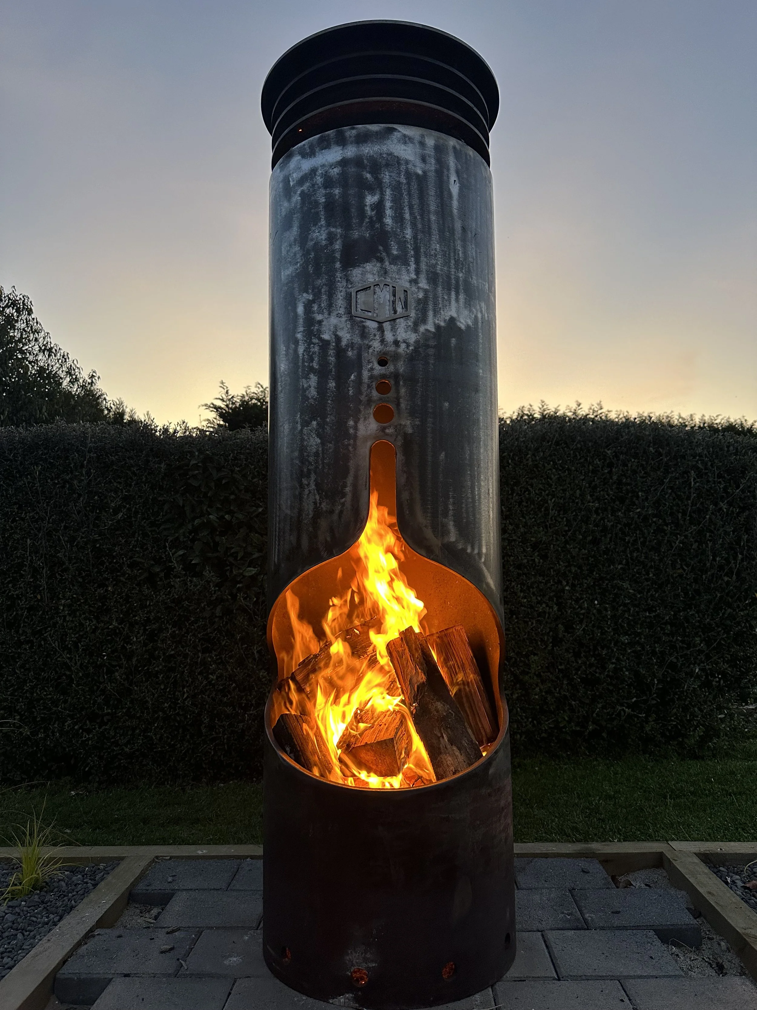 Outdoor wood-burning chiminea with a tall metal chimney, glowing with a bright fire inside, set on a stone patio with a hedge and garden in the background.