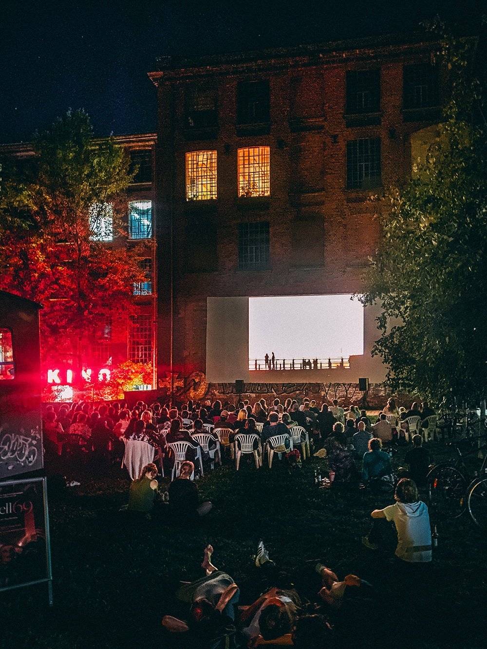 Open-Air-Kino-Veranstaltung in einem Hinterhof bei Nacht, Menschen sitzen auf Plastikstühlen vor einer großen Leinwand, Gebäude im Hintergrund beleuchtet.