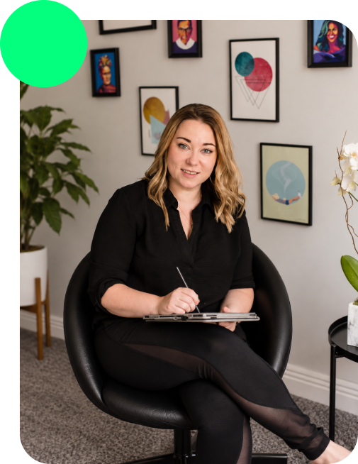 A woman sitting in a black chair, holding a notebook and pen, in a room with framed colorful artwork on the wall and a potted plant nearby.