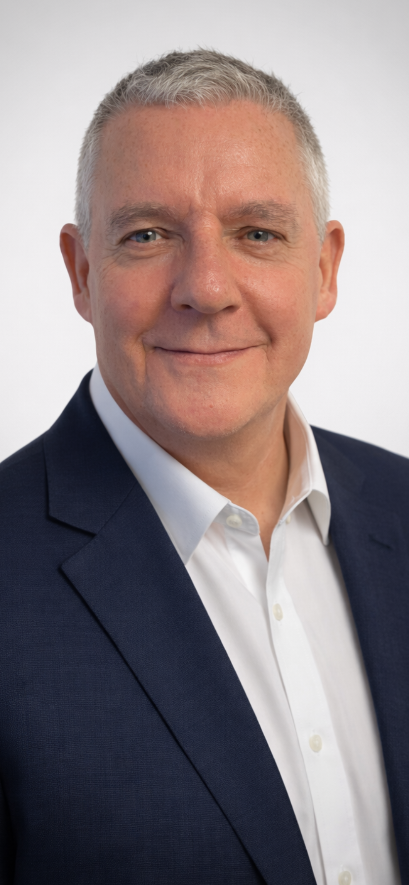 Headshot of a middle-aged man with short gray hair, blue eyes, smiling, dressed in a white collared shirt and dark blazer, on a plain light background.