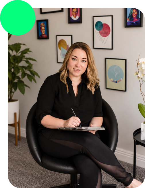 A young woman with blonde, wavy hair and a black shirt sitting in a black armchair, holding a clipboard and pen, in a room decorated with framed artwork on the wall and potted plants.