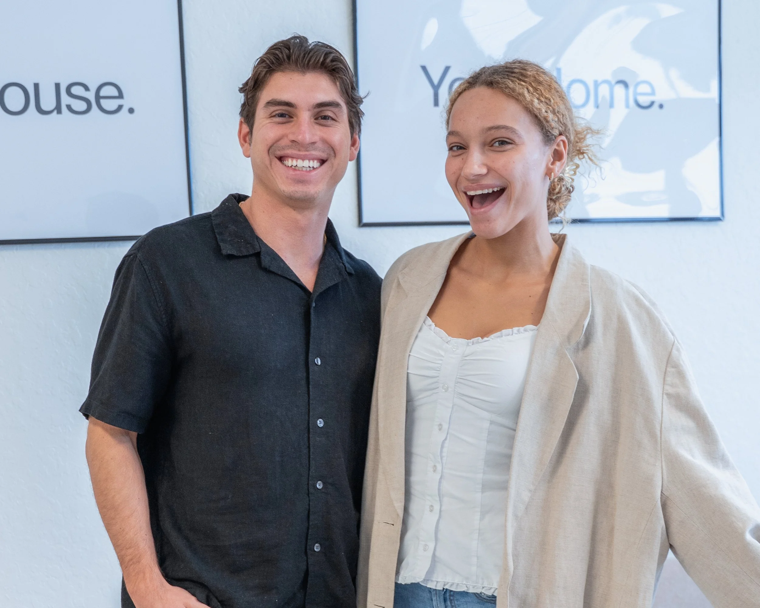 A smiling man and woman standing together indoors with a white wall and a framed picture in the background.