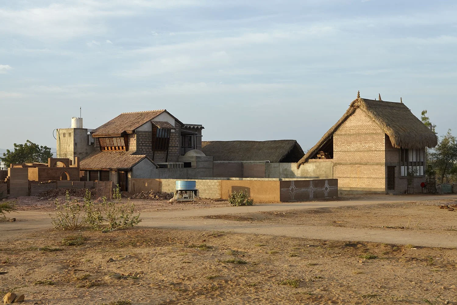 Rural scene with traditional brick and thatched-roof houses in a dry area with sparse vegetation and a dirt road in front, under a cloudy sky.