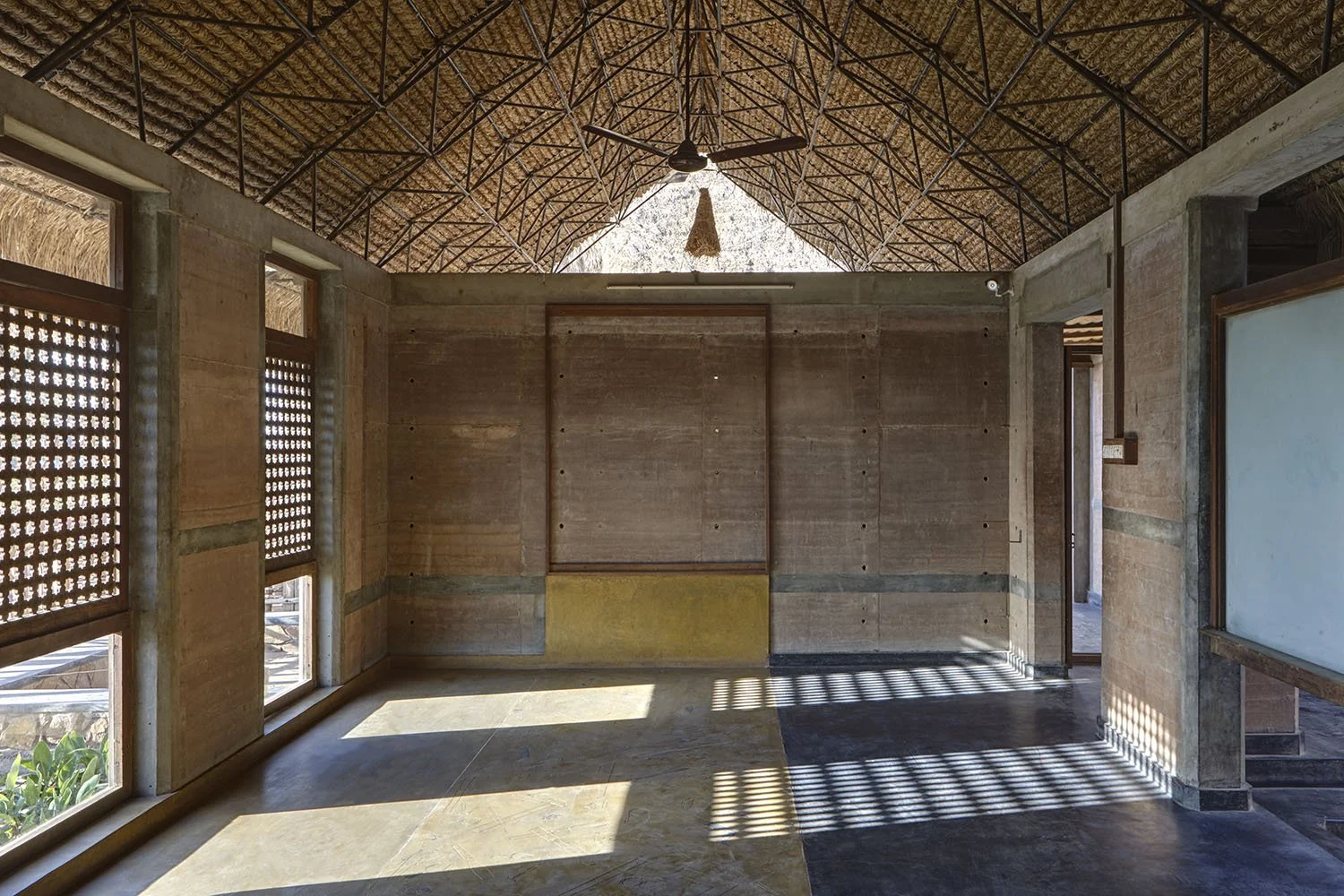 Interior of a building with a thatched roof, concrete walls, and large windows with wooden lattice panels, allowing sunlight and shadows to create patterns on the floor.