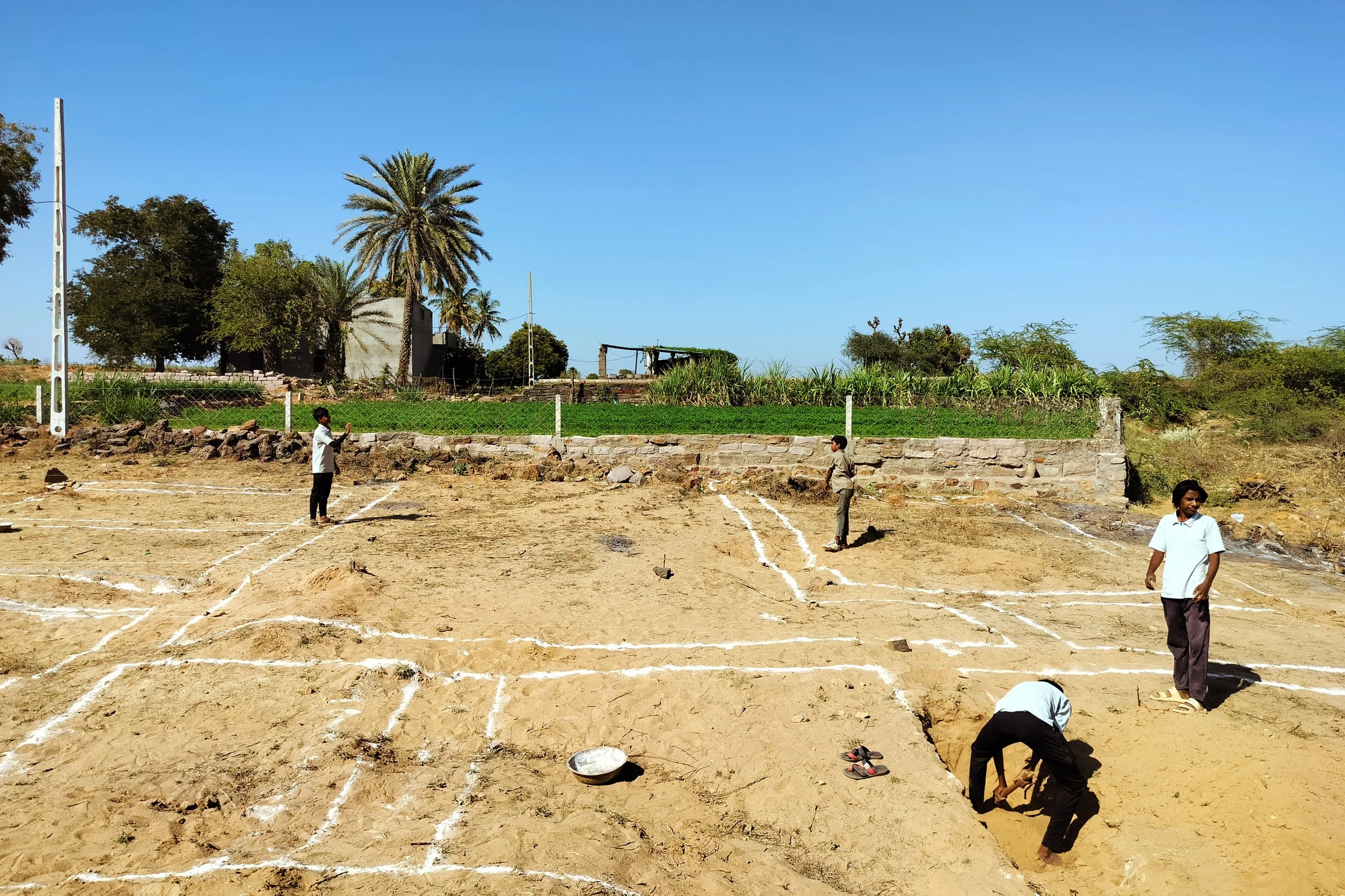 People marking a sports field with white paint on dry soil, with trees and a clear blue sky in the background.