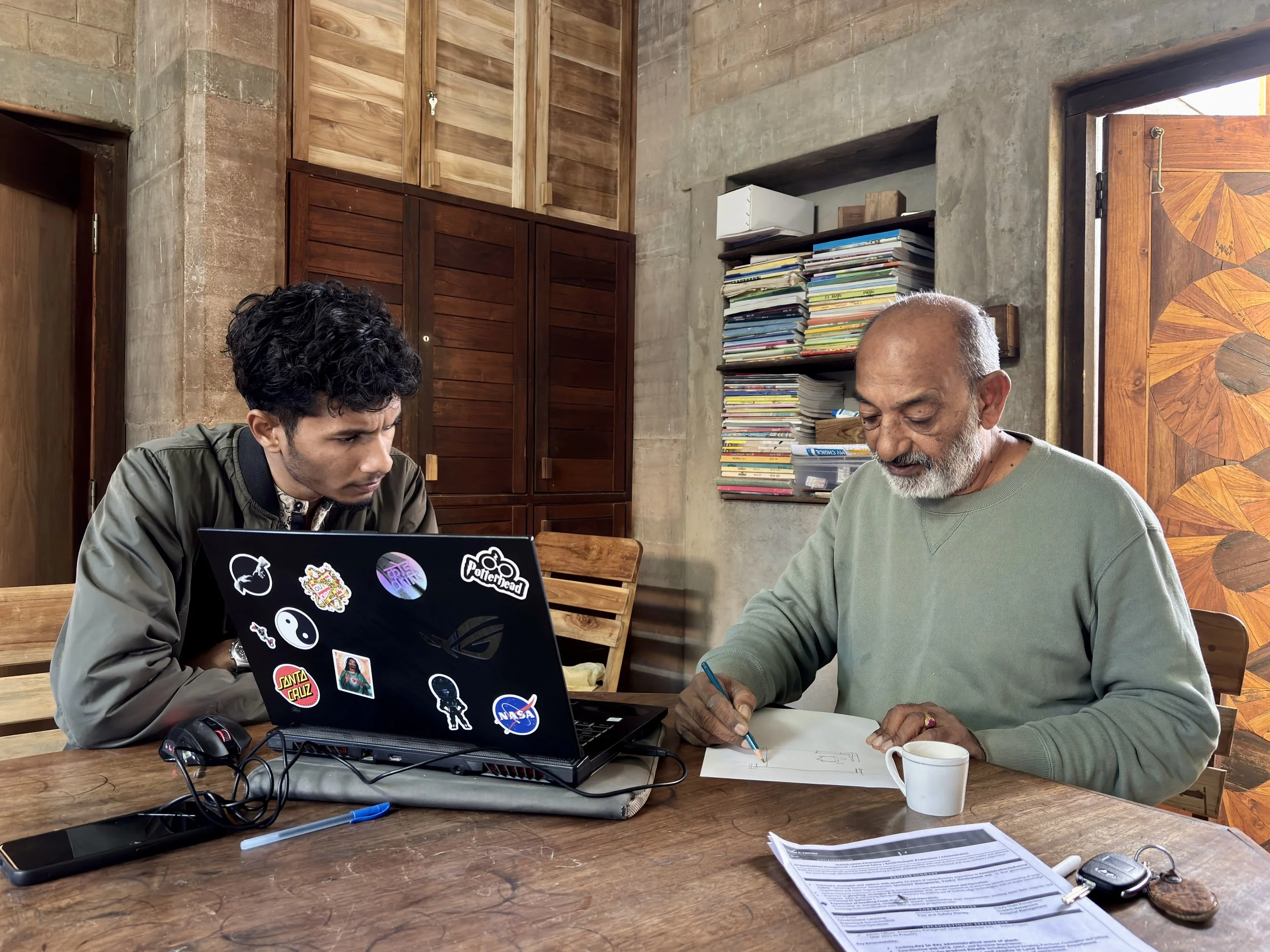 Two men sitting at a wooden table, one young with black curly hair working on a laptop with stickers, the other older with gray hair and beard writing on paper with a cup of coffee nearby, in a room with wooden and concrete walls and a shelf full of stacked papers or books.