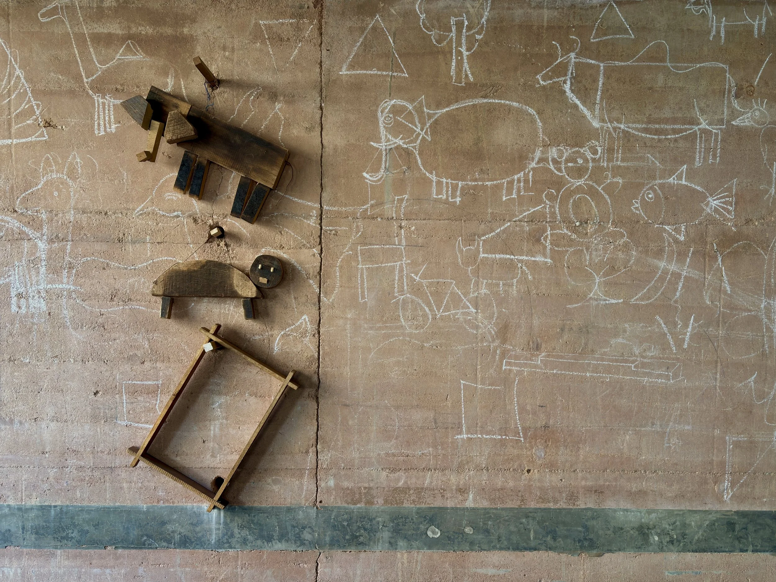 A concrete wall with chalk drawings of animals and shapes. There are wooden toy animals and a wooden frame on the floor.