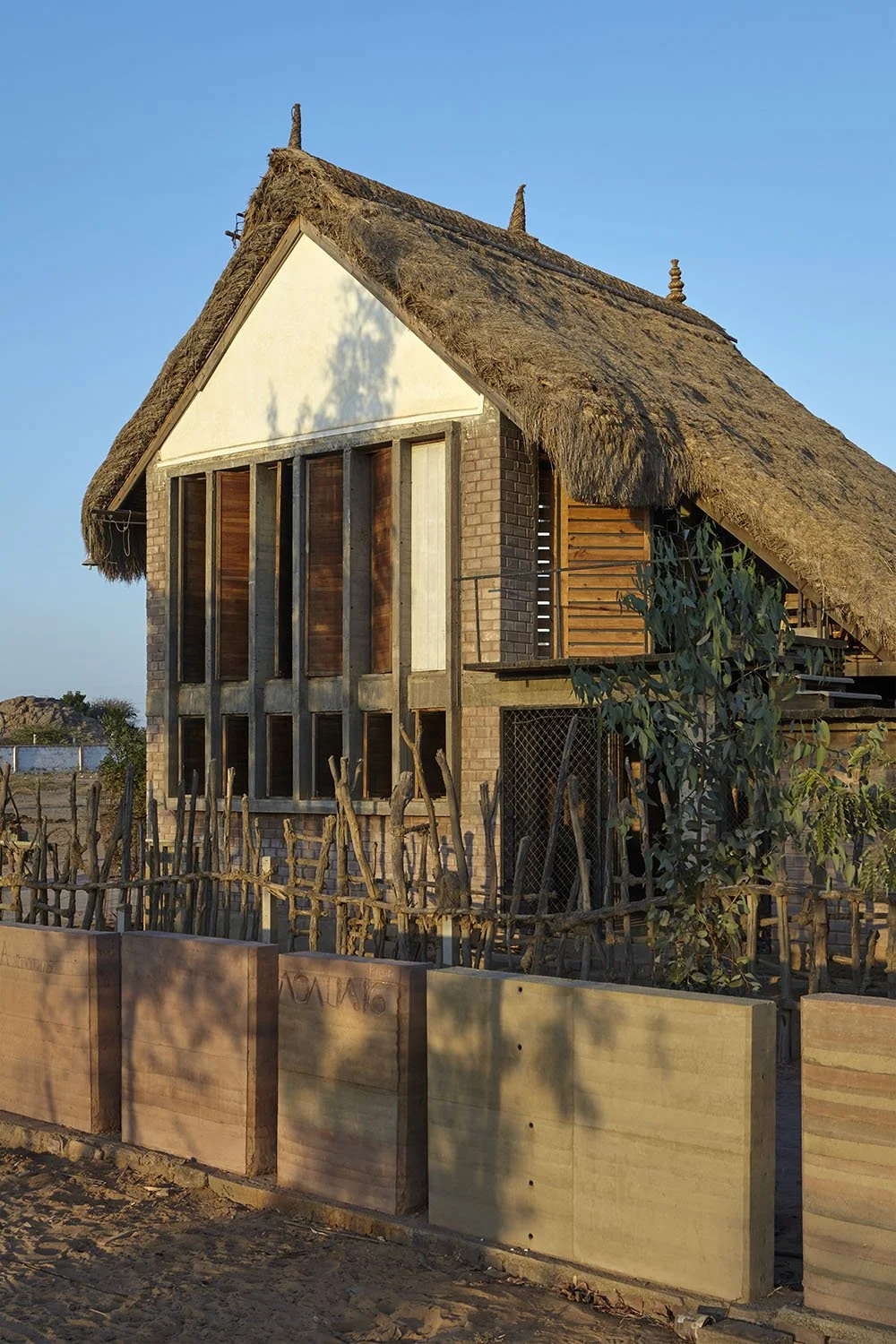 A house with a thatched roof, brick and wooden walls, and a small enclosed yard, under a clear blue sky.