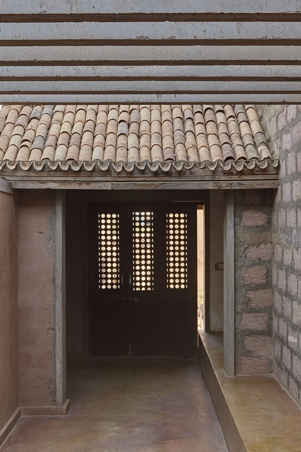 A rustic outdoor entrance area with a tiled roof, brick walls, and a wooden door with decorative lattice panels, illuminated by natural sunlight.