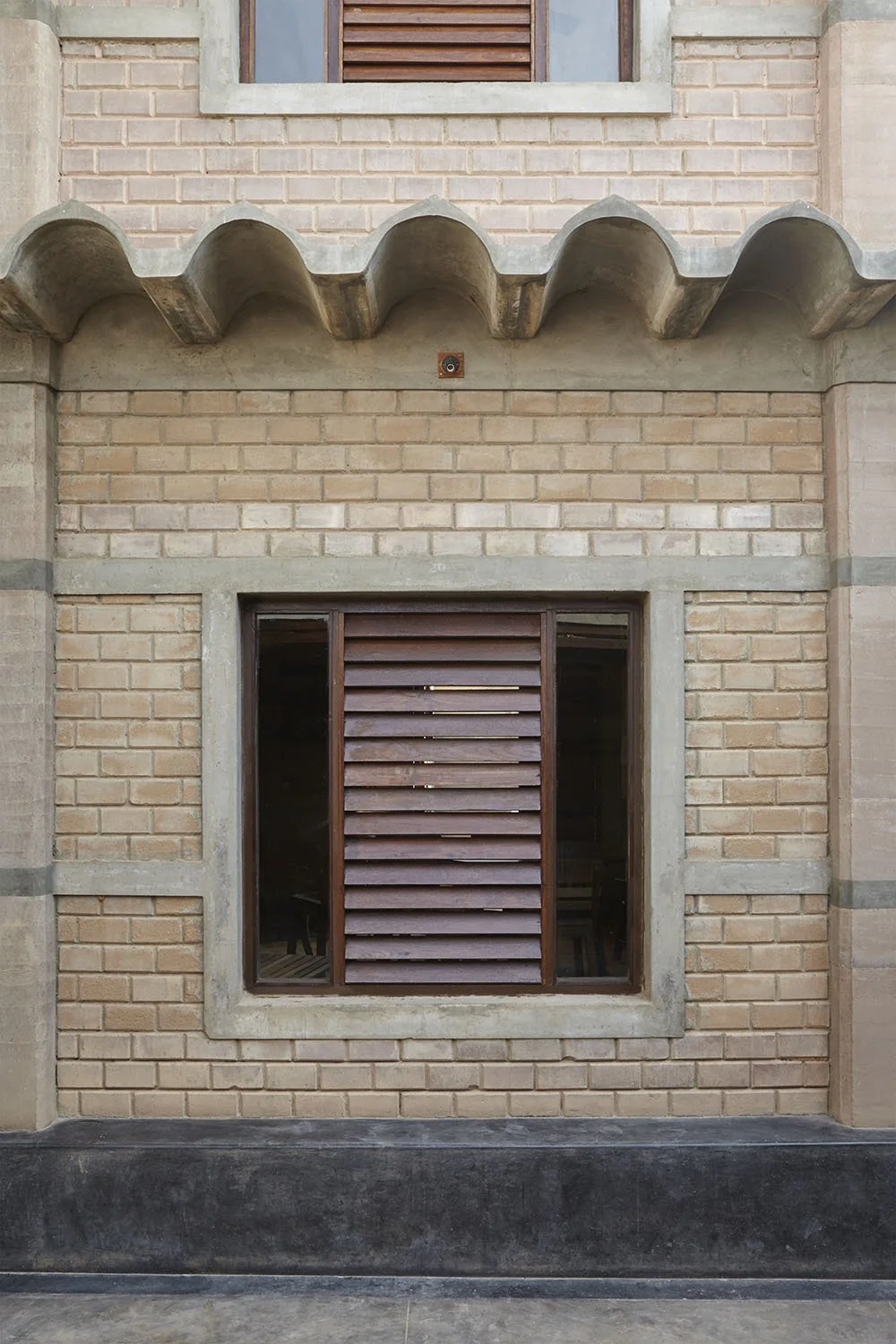 A brick building with a window partially covered by wooden shutters, above a decorative concrete arch, with a small round ventilation opening near the top.