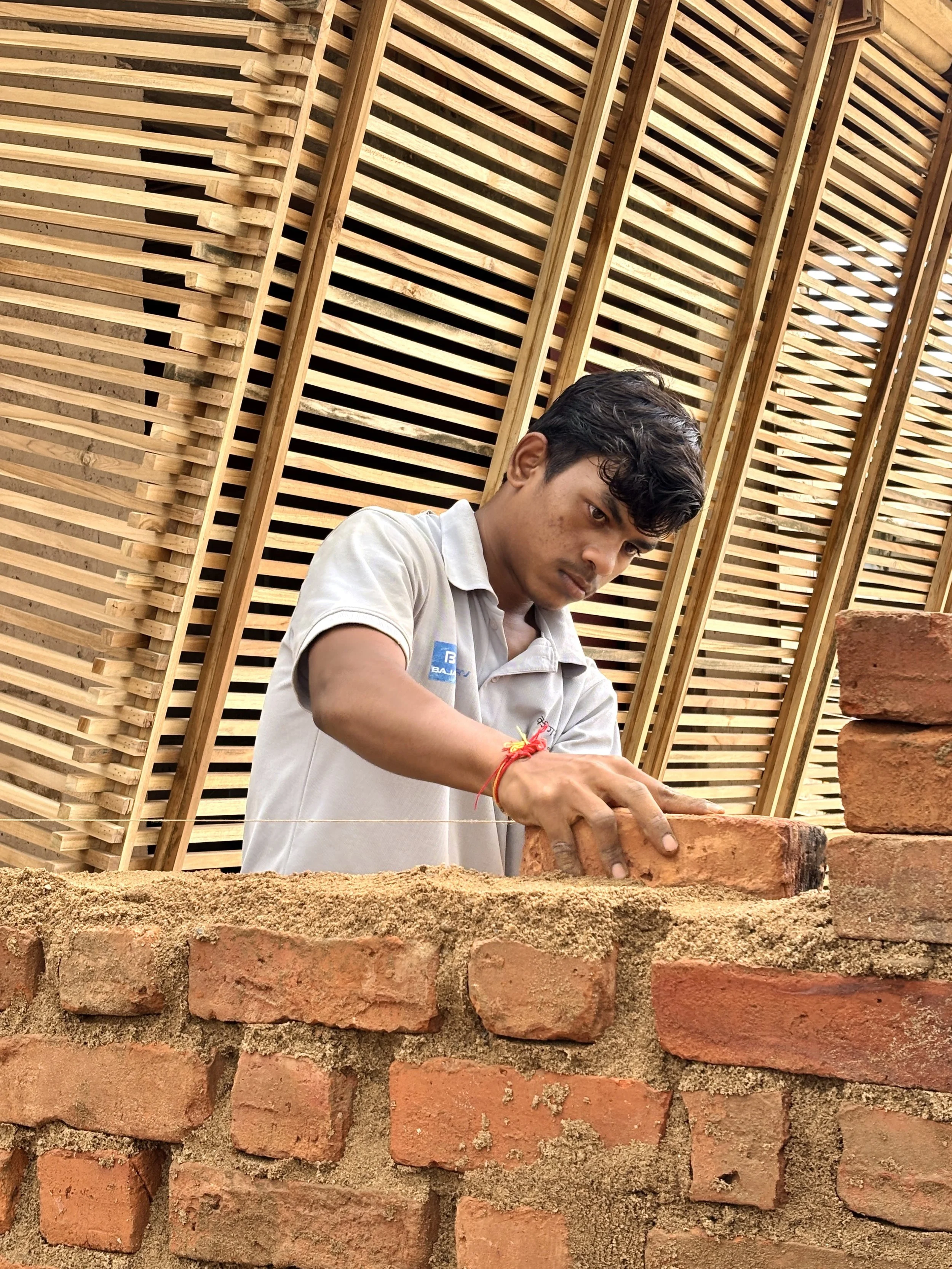A young man building a brick wall, placing a brick on top of the wall, against a backdrop of wooden slats arranged vertically.