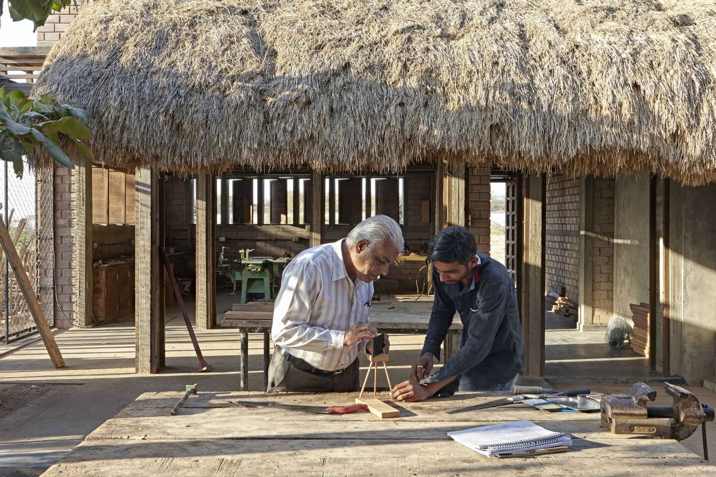 Two men working together on a woodworking project outdoors under a thatched roof. One older man and one younger man are measuring a piece of wood on a workbench surrounded by tools and plans.