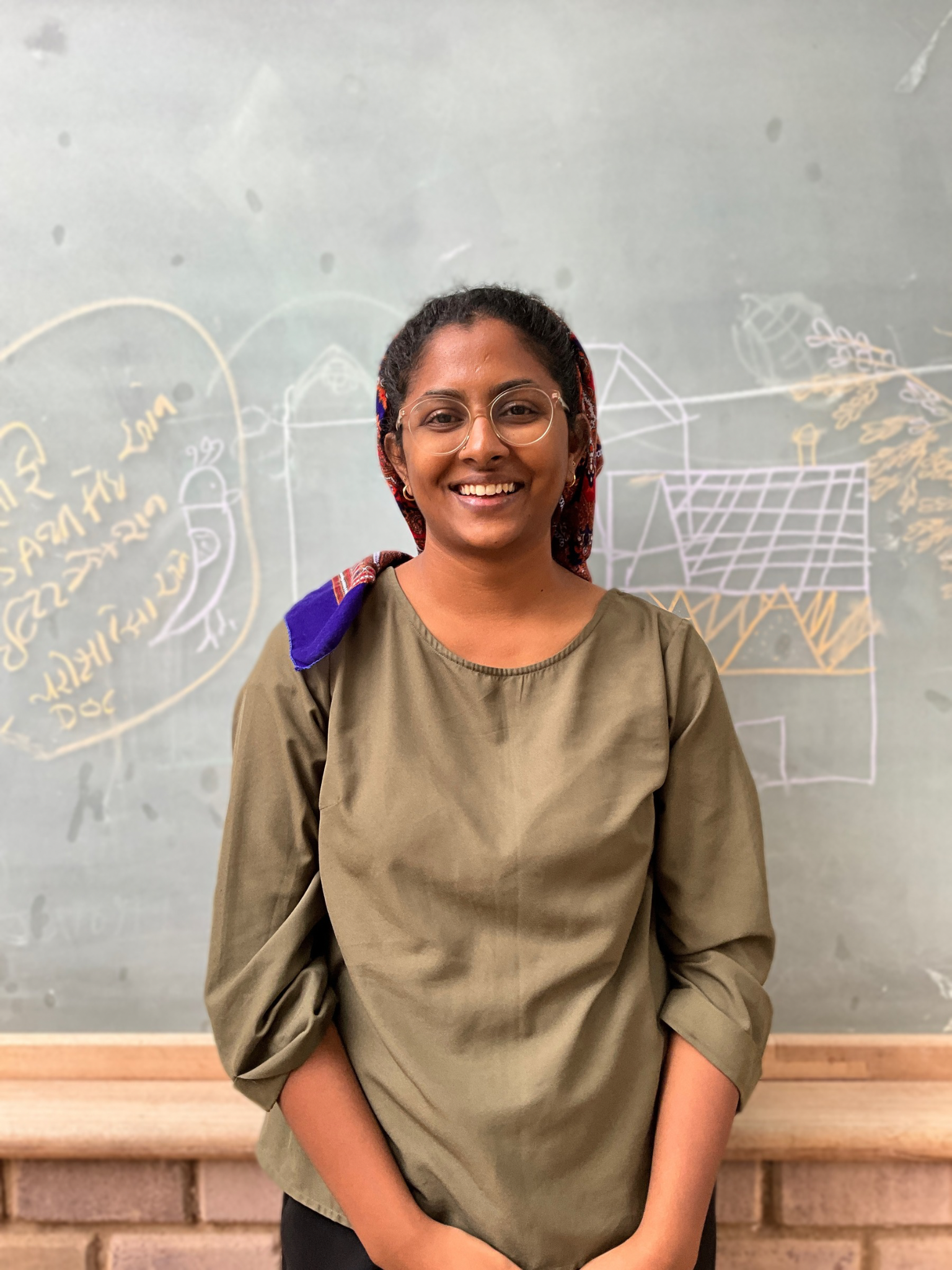 A smiling woman with glasses and a colorful headscarf standing in front of a chalkboard with drawings and writing.