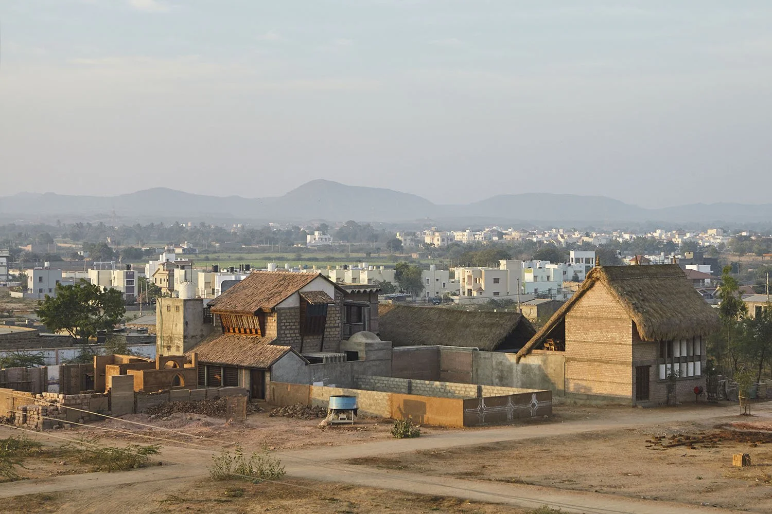 A rural area with traditional houses having thatched roofs and brick walls, set against a backdrop of modern white buildings and distant mountains.