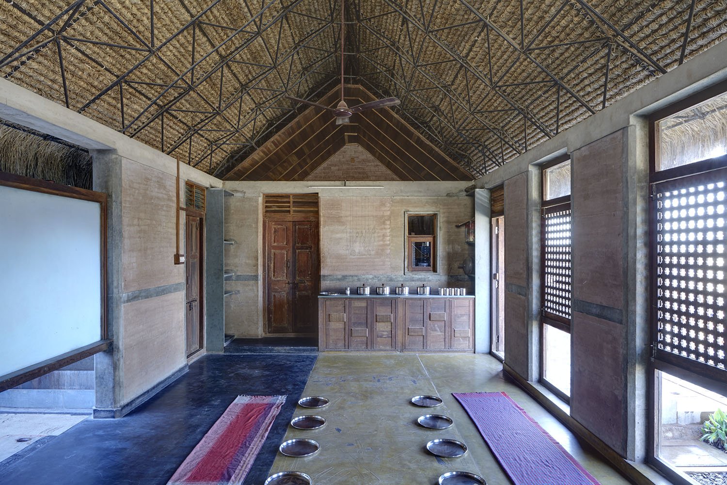 Interior of a rustic, open-concept room with a high ceiling made of thatched roof and wooden beams. There are large windows with wooden shutters on the right side. The floor has a mix of dark and light tiles, with two red mats. Several small metal pl