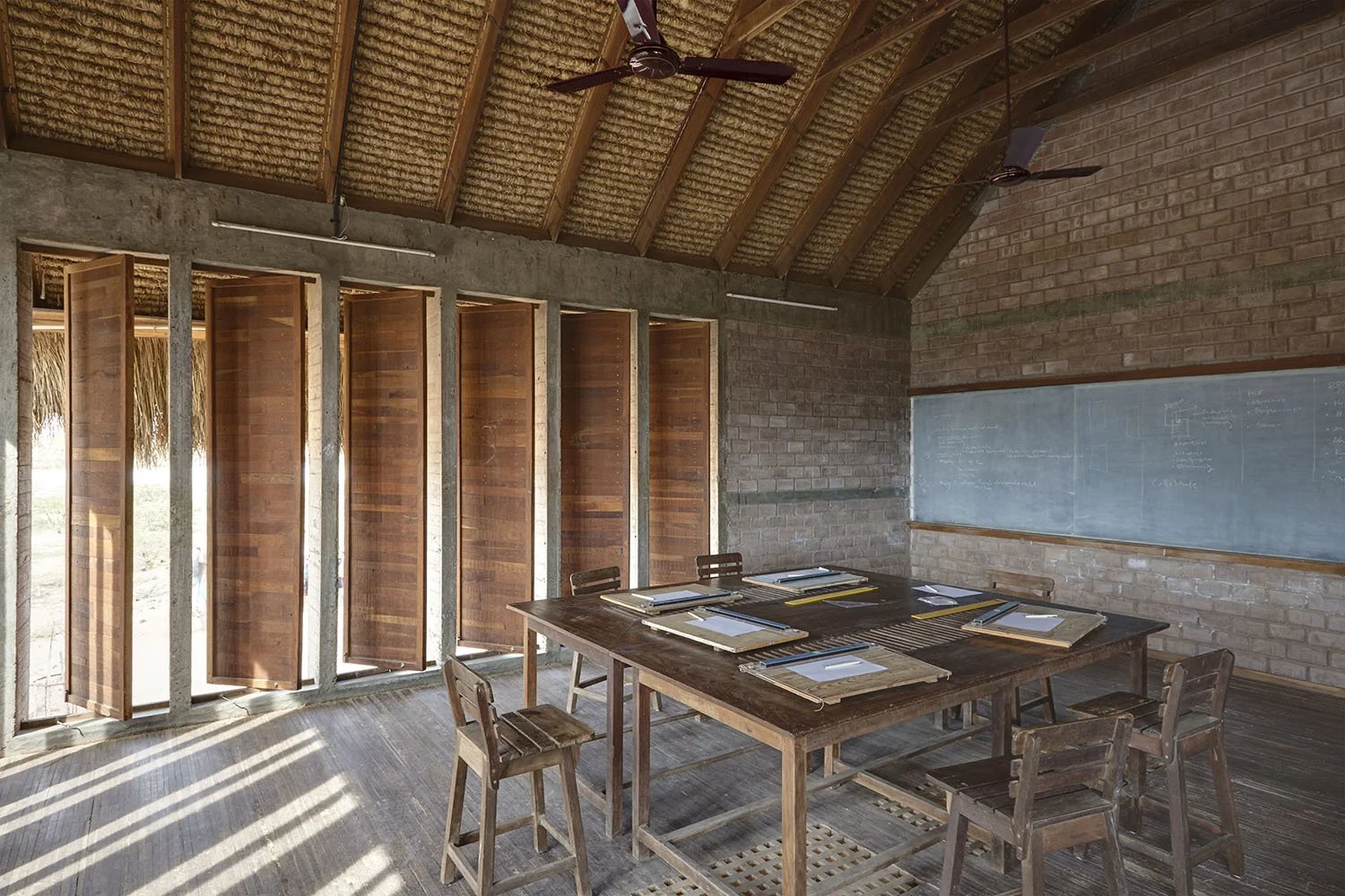 Interior of a classroom or meeting room with a wooden table, chairs, a chalkboard, and a ceiling fan.