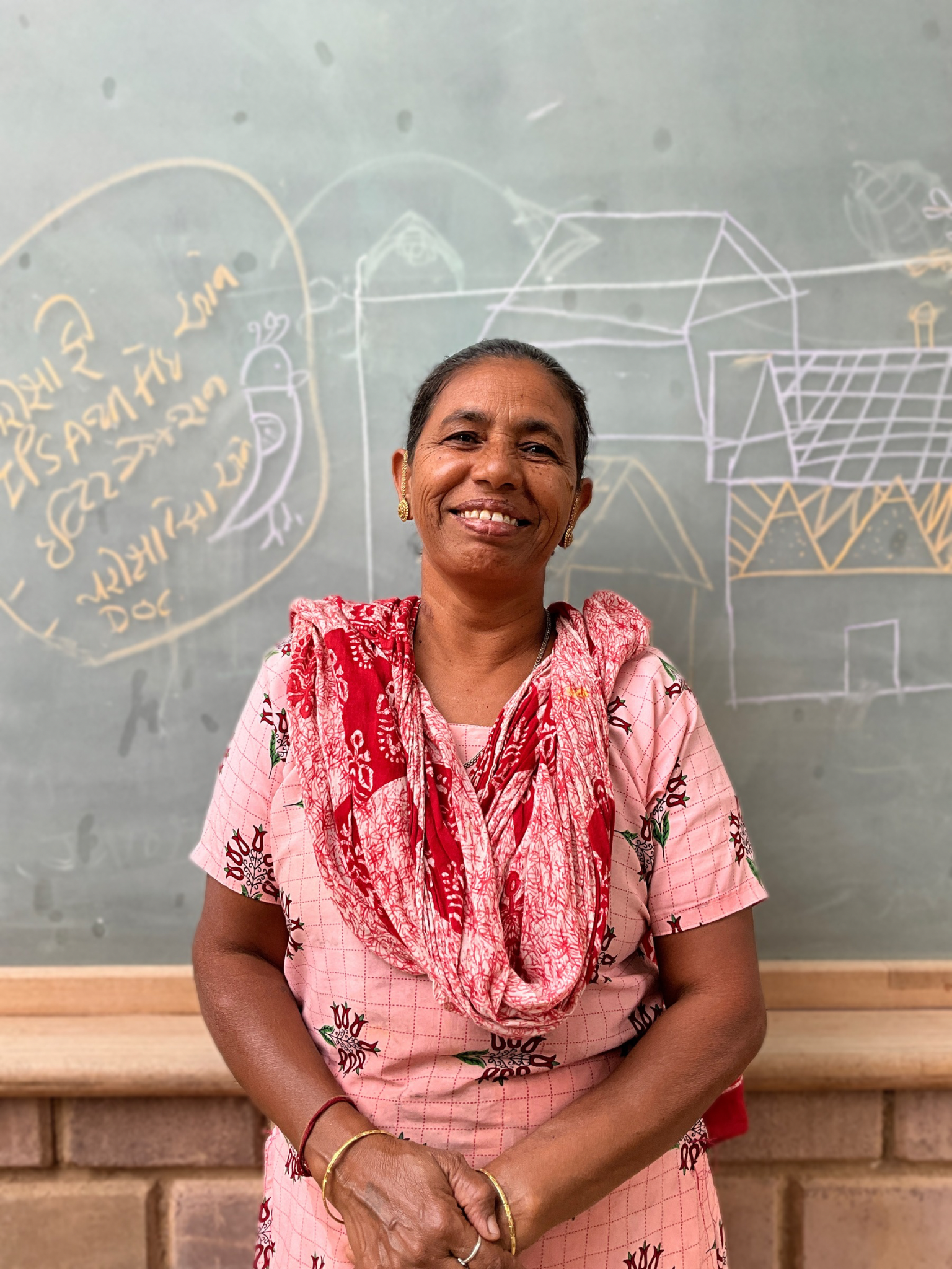 A smiling woman standing in front of a chalkboard with colorful sketches and writings, wearing a pink patterned dress and a red scarf.