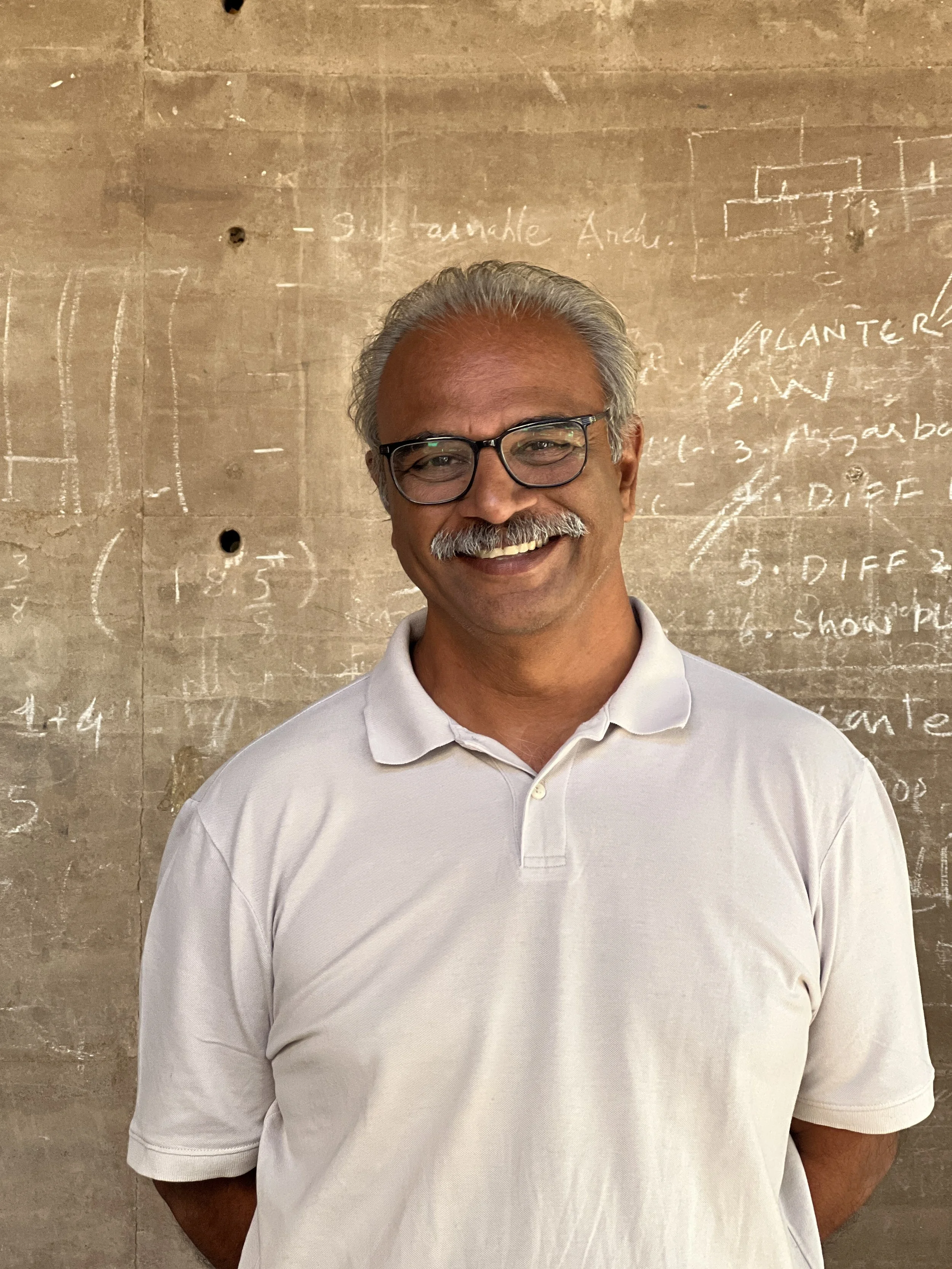 A smiling man with gray hair and a mustache wearing glasses and a white polo shirt standing in front of a wall with chalk writing and diagrams.
