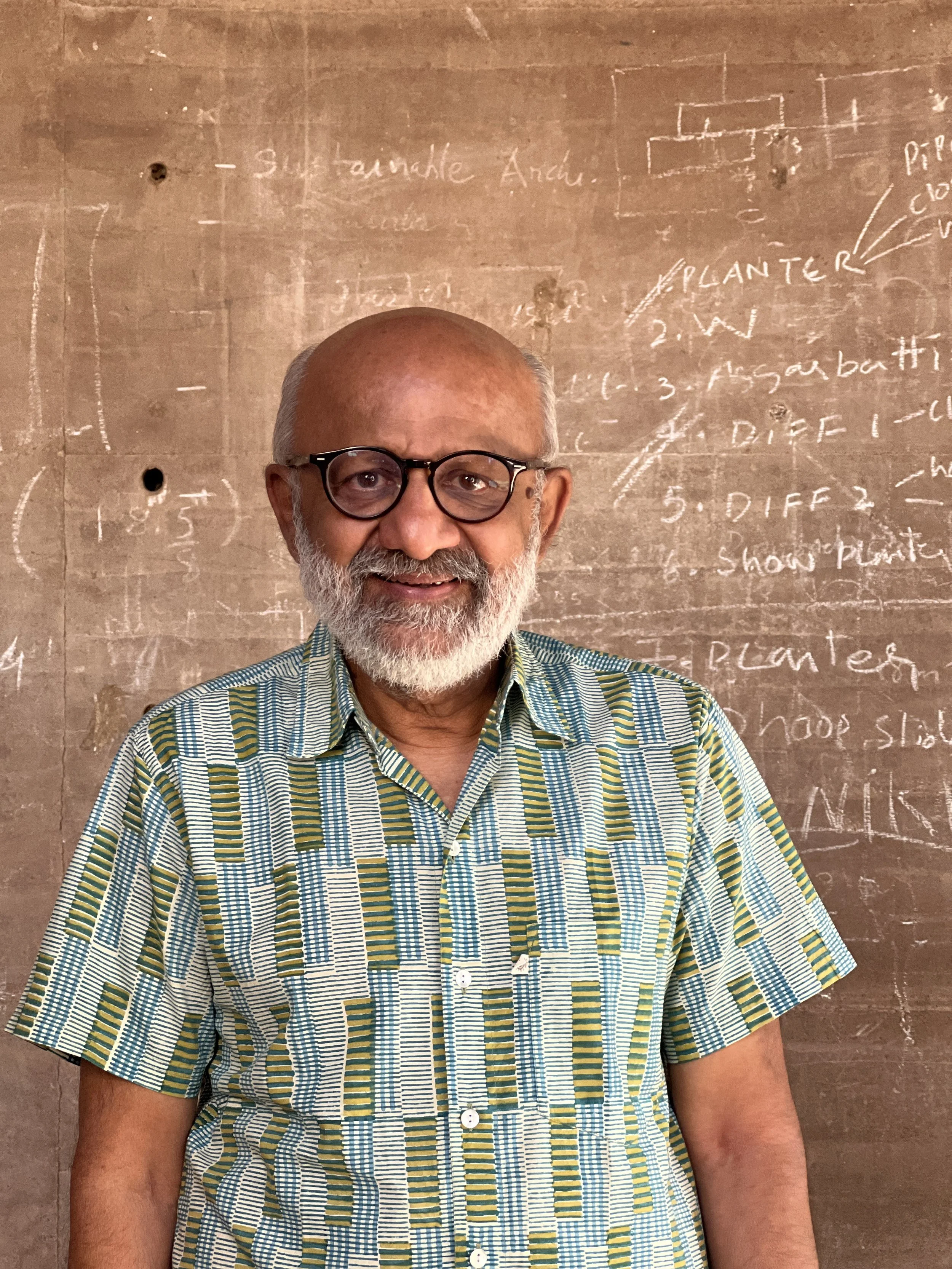 An elderly man with glasses and a beard, smiling, standing in front of a chalkboard with written notes and diagrams.